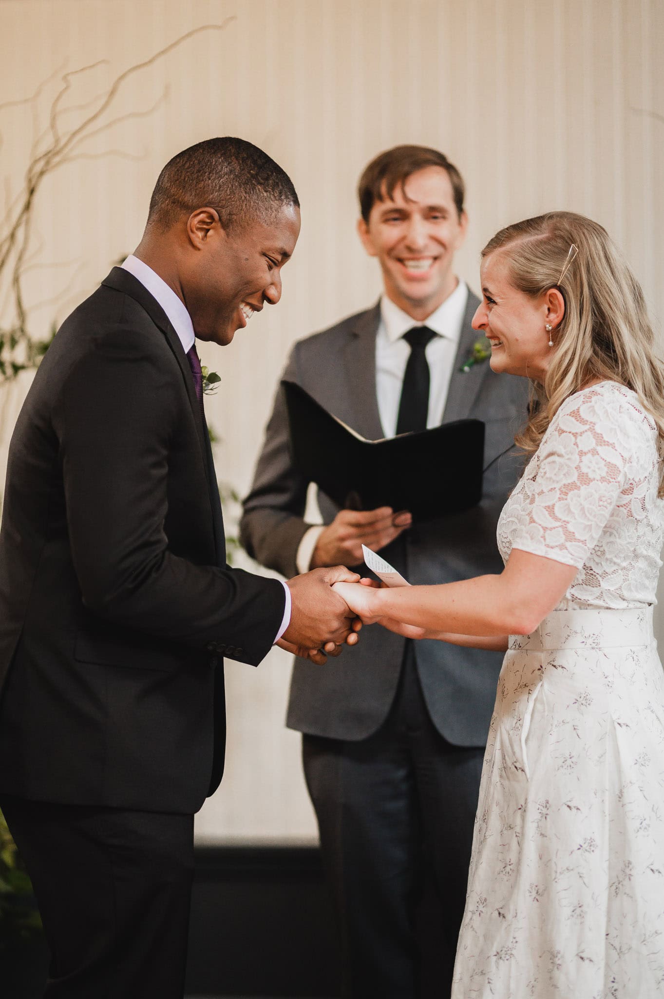 A couple exchanging vows, smiling, with a person officiating the wedding ceremony in the background at the enchanting wedding at the Evergreen.