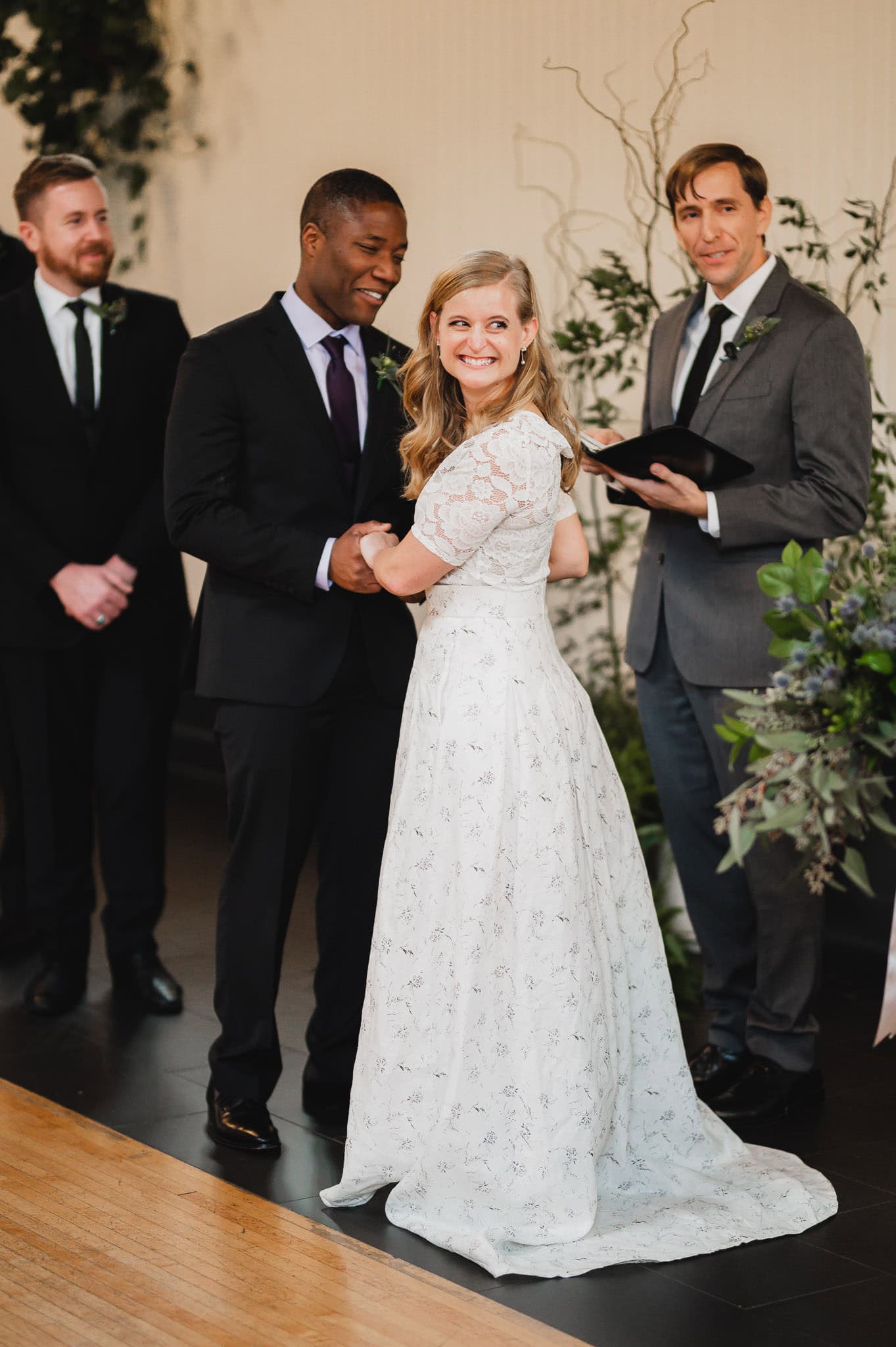 During their wedding at the Evergreen, a bride and groom hold hands, with a man officiating and another standing nearby.