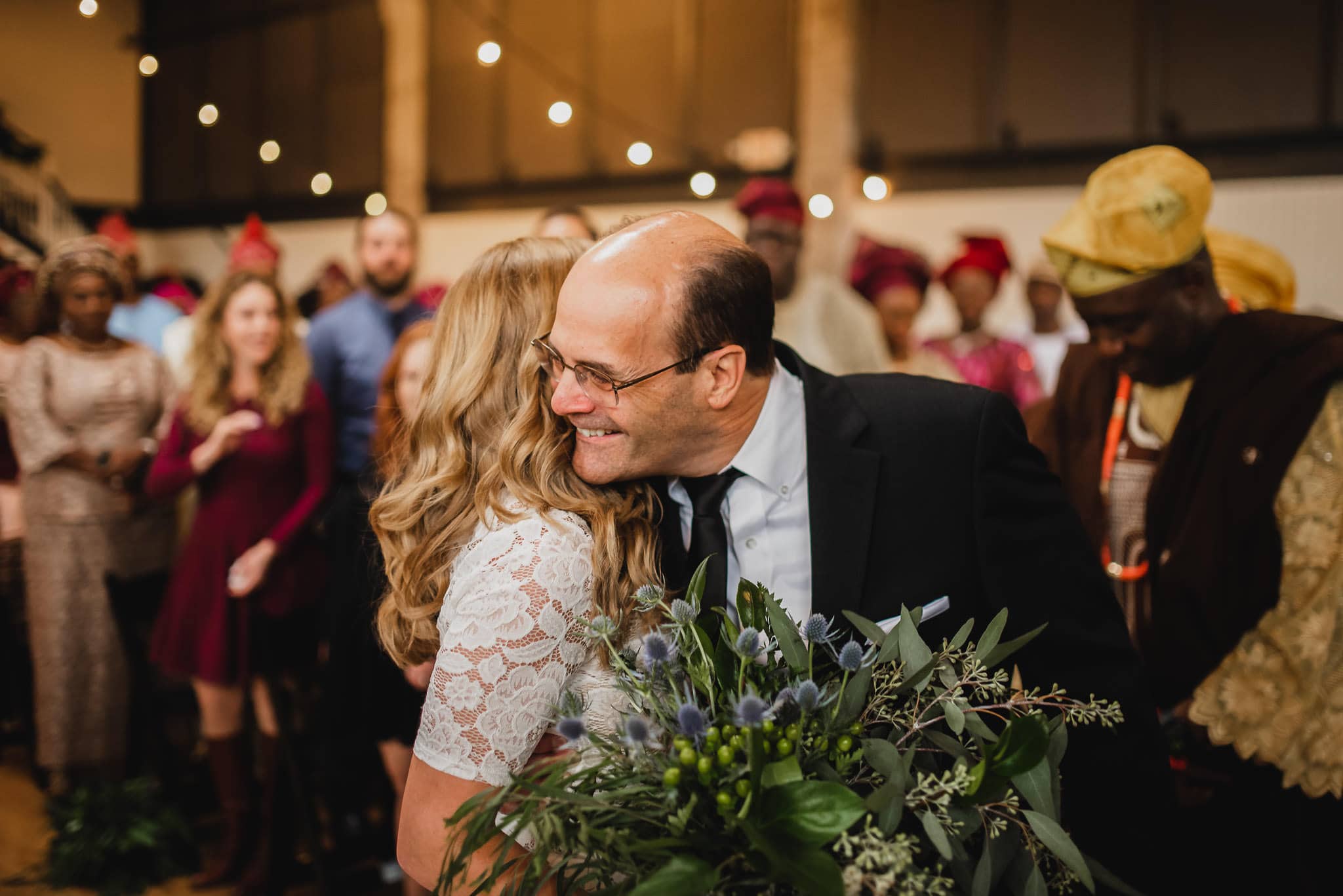 At the wedding at the Evergreen, a man in a suit embraces a woman holding a bouquet during an indoor gathering. People dressed in colorful attire are visible in the background.