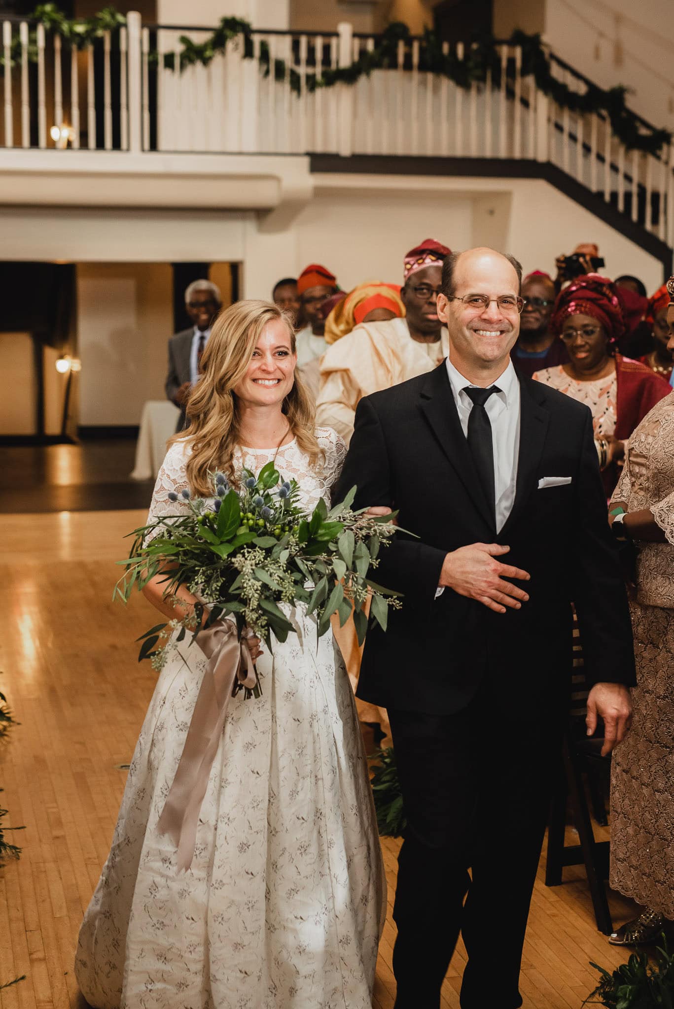 The bride and man in a suit smile as they walk down the aisle at the Evergreen. Guests in colorful attire stand in the background, adding to the joyous atmosphere.