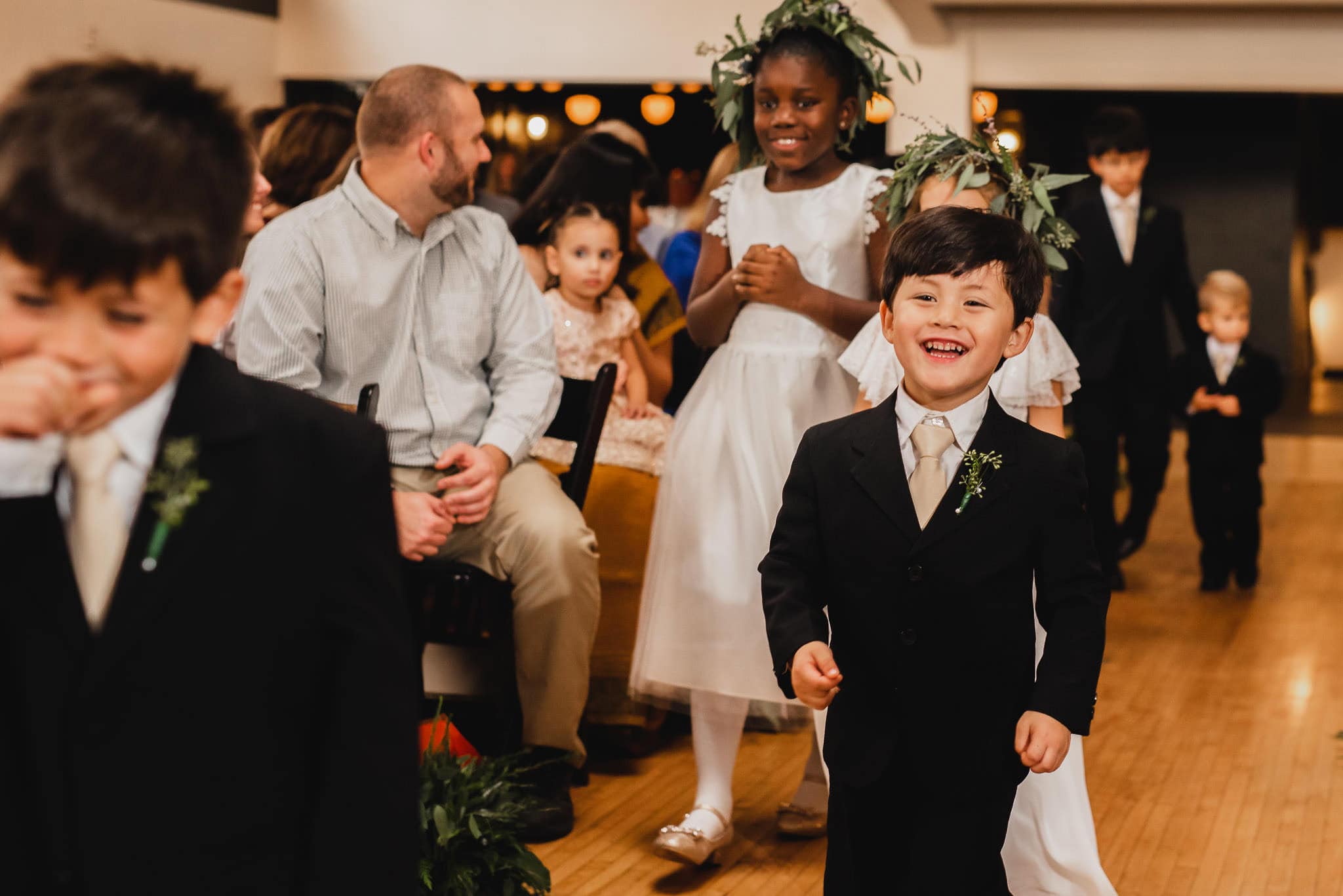 Children dressed in formal attire walk down the aisle at a wedding at the Evergreen, smiling and surrounded by seated guests.