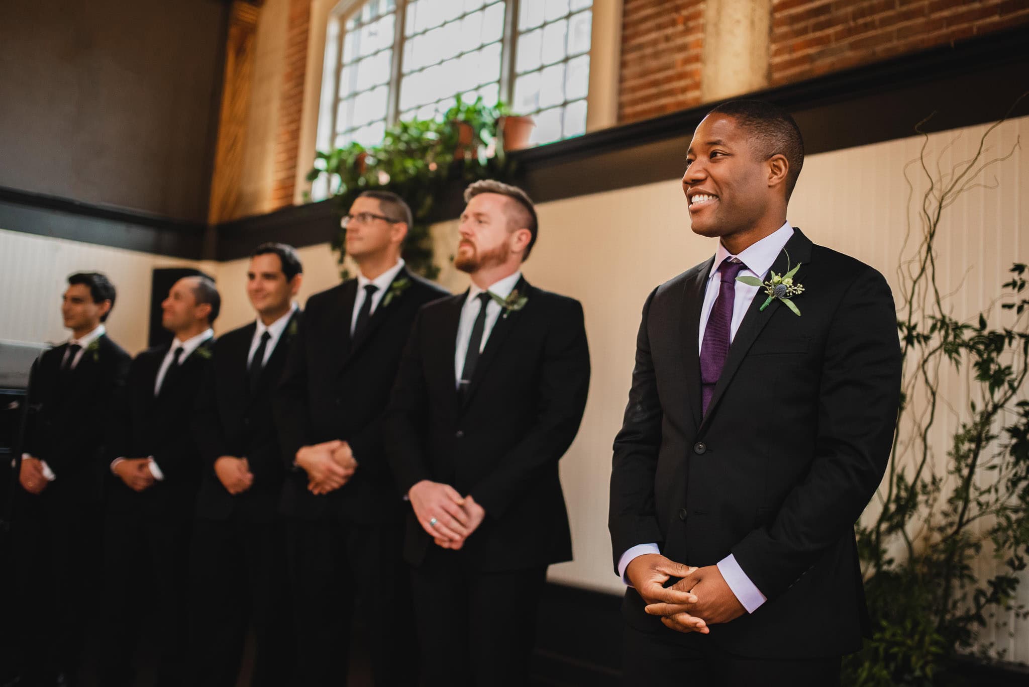 A group of groomsmen in suits stand indoors at the wedding at the Evergreen, smiling and lined up, with plants and a window in the background.