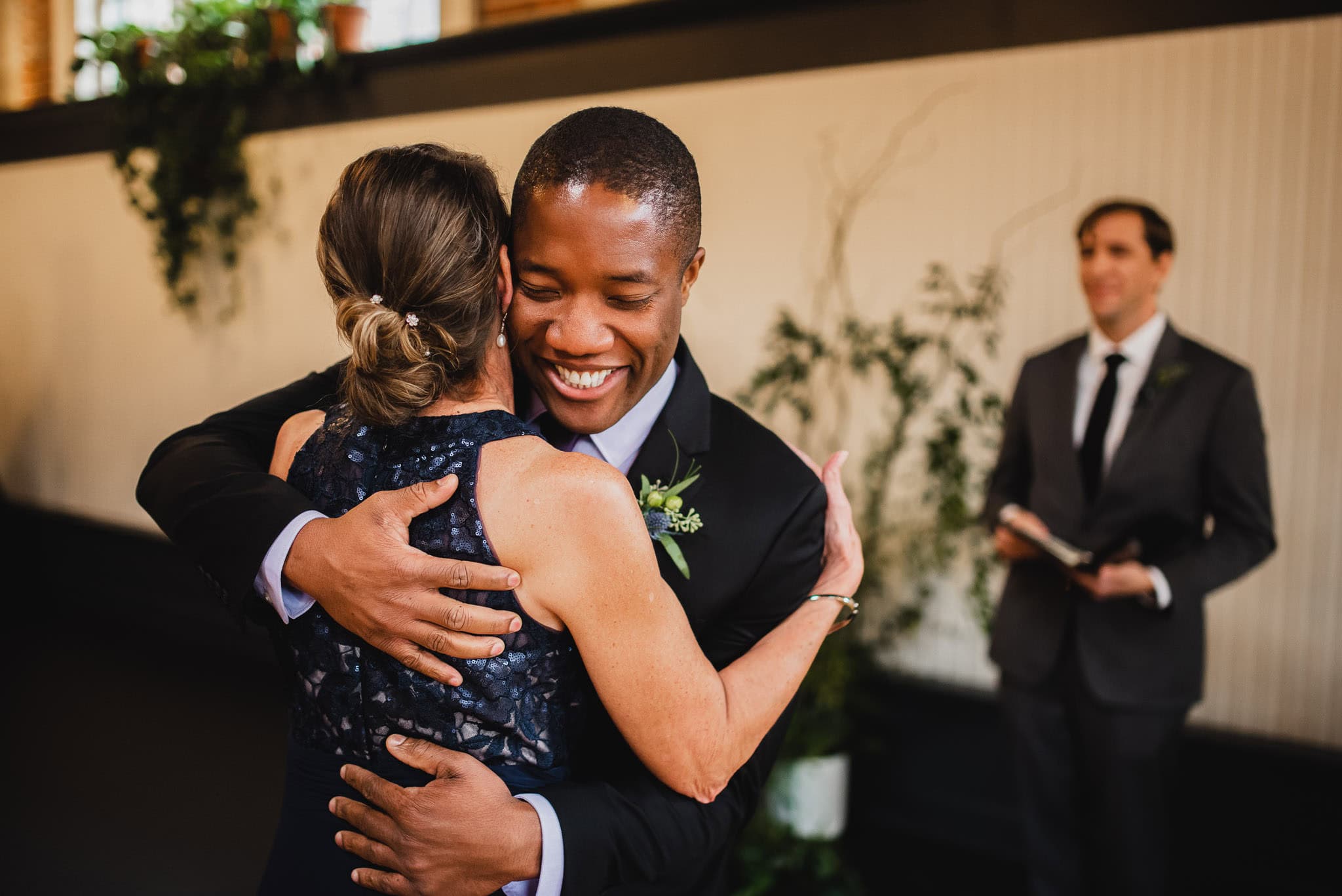 A smiling person in formal attire hugs a woman in a dark dress, with a man in a suit observing in the background, capturing the joyous atmosphere of a wedding at the Evergreen.