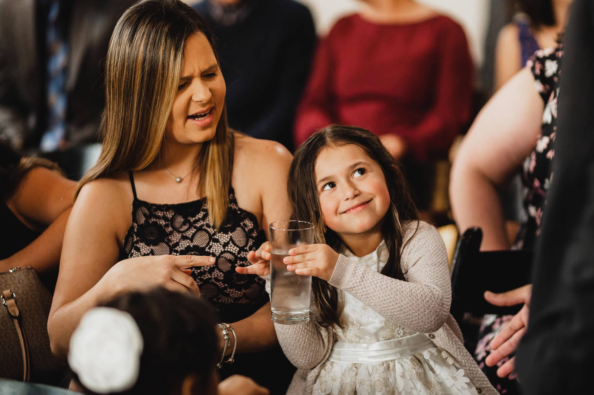 At the wedding at the Evergreen, a woman and girl are smiling and sitting among guests. The girl holds a glass of water, capturing a moment of pure joy in the celebration.