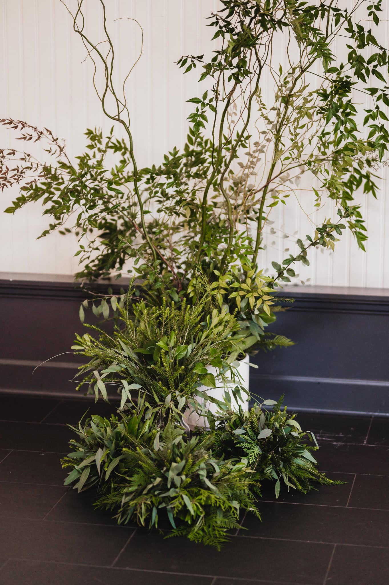 Lush green plants arranged in a tall white pot, set against an elegant white and black wall backdrop, perfect for a wedding at the Evergreen.