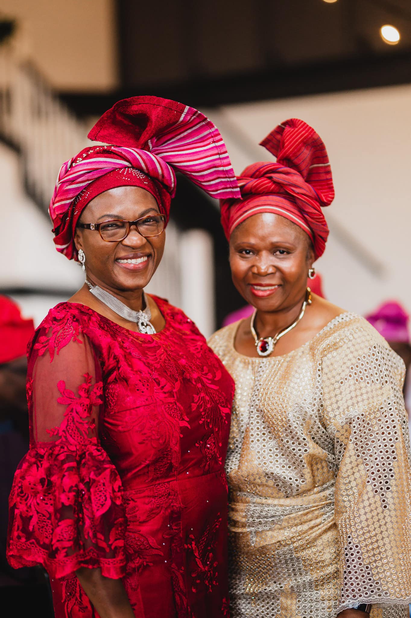Two women in elegant traditional attire and red headwraps are smiling indoors, possibly preparing for a wedding at the Evergreen.