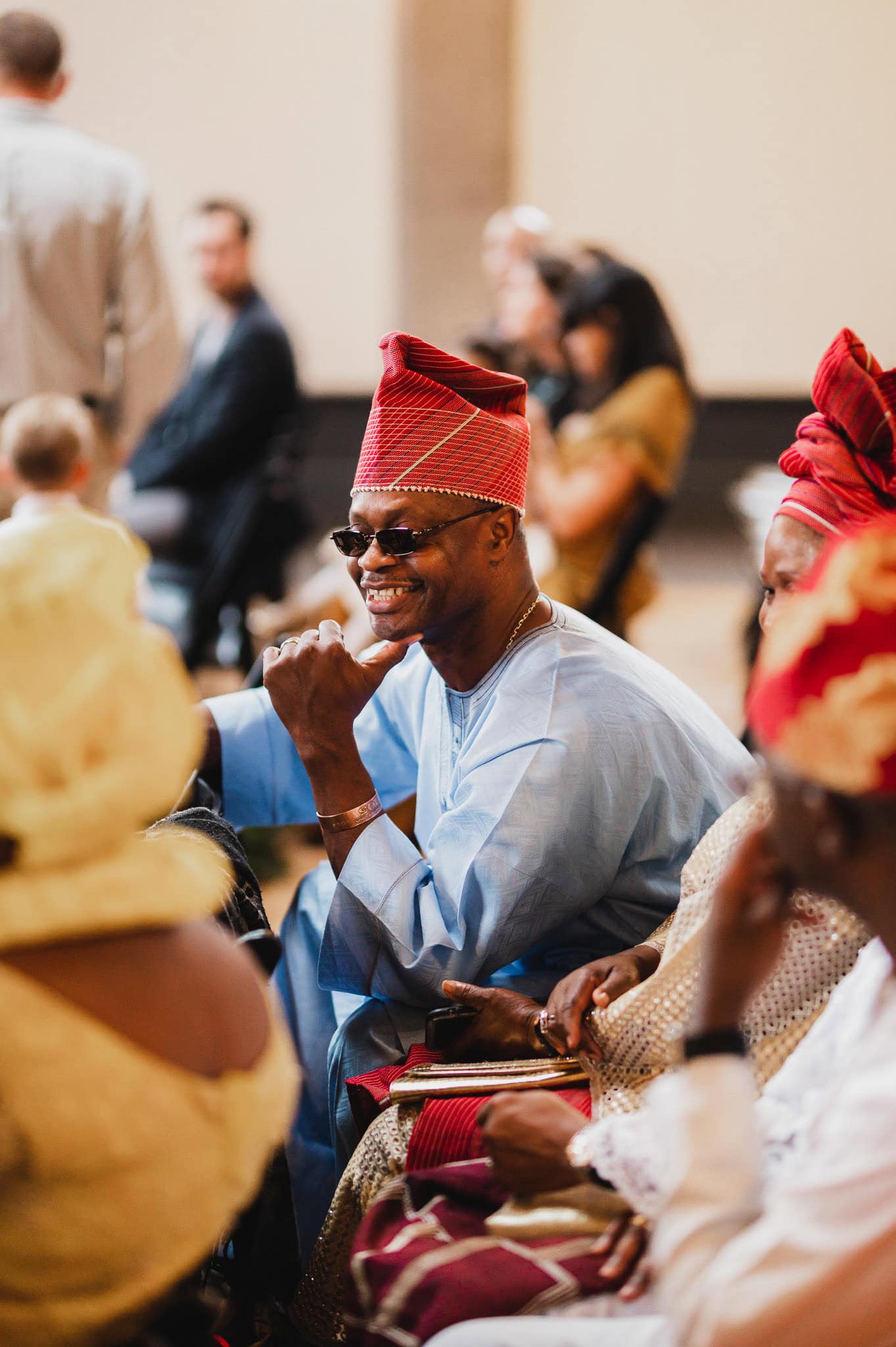 A man in blue attire and a red cap is smiling at a wedding at the Evergreen, surrounded by others in colorful traditional clothing.