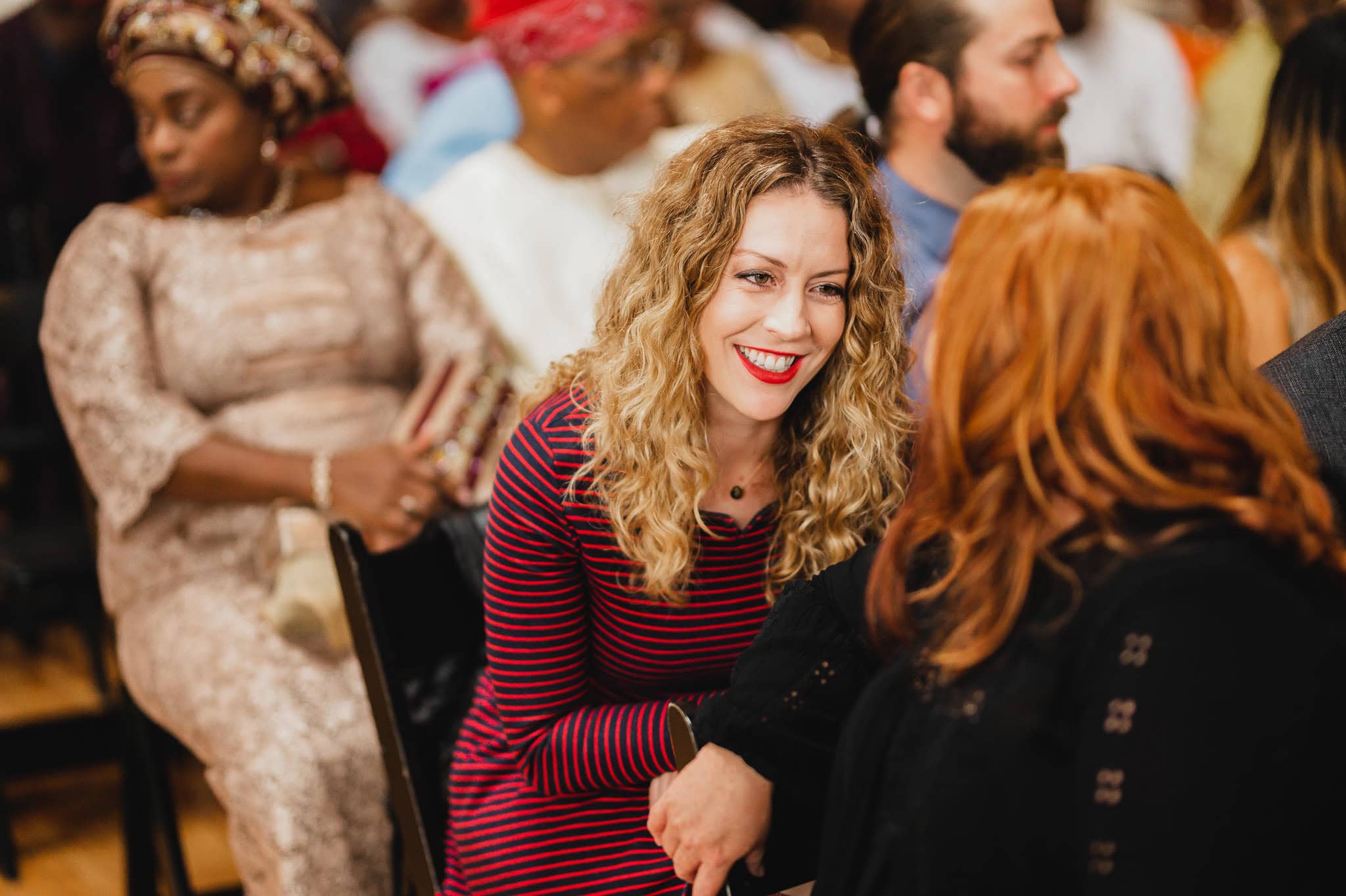 At a bustling indoor event, a woman with curly hair and a striped dress smiles and chats warmly with someone. It feels like she could be enjoying herself at a wedding at the Evergreen.