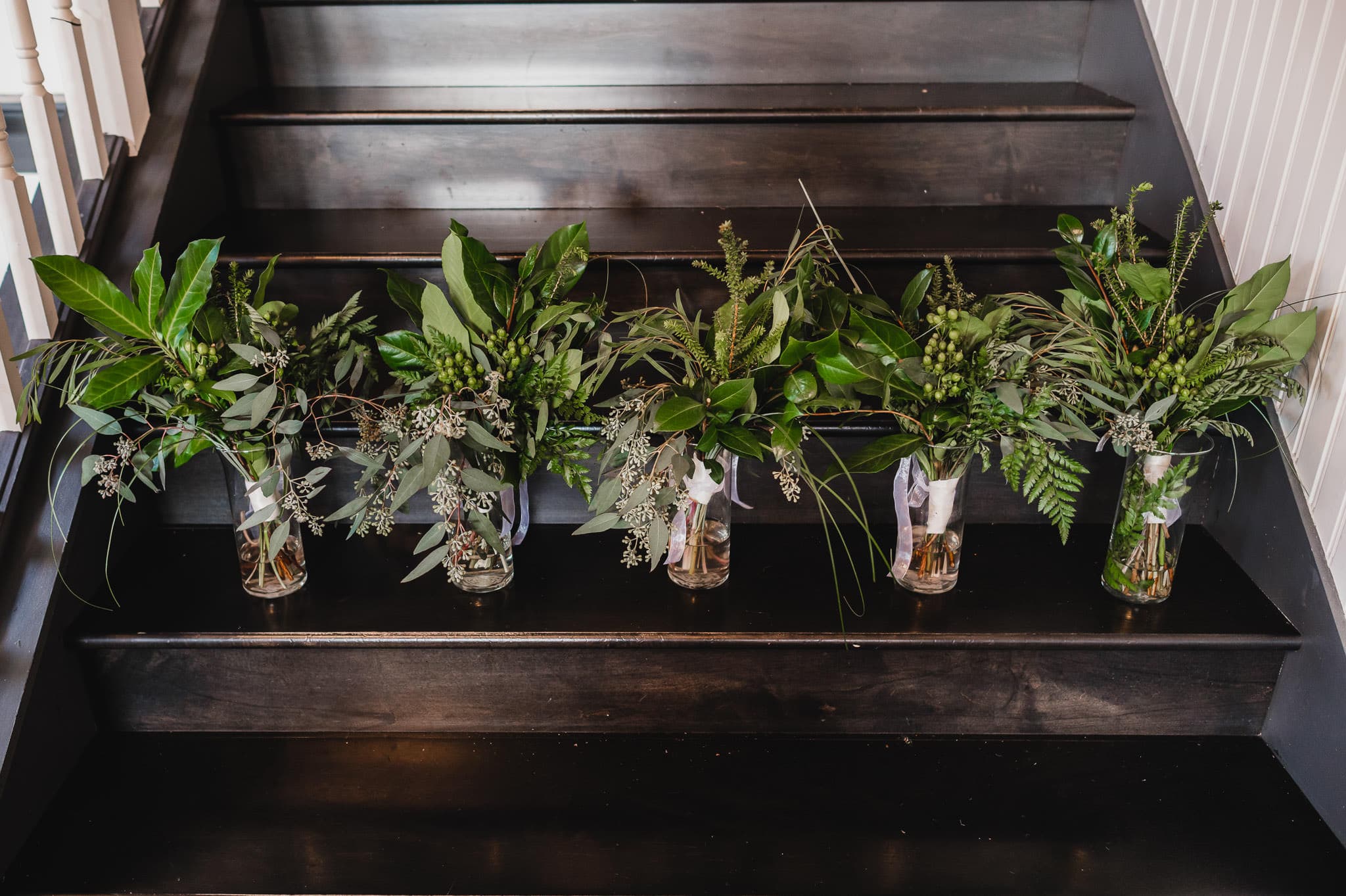 Five glass vases with green foliage elegantly arranged on dark wooden stairs set the scene for a beautiful wedding at the Evergreen.