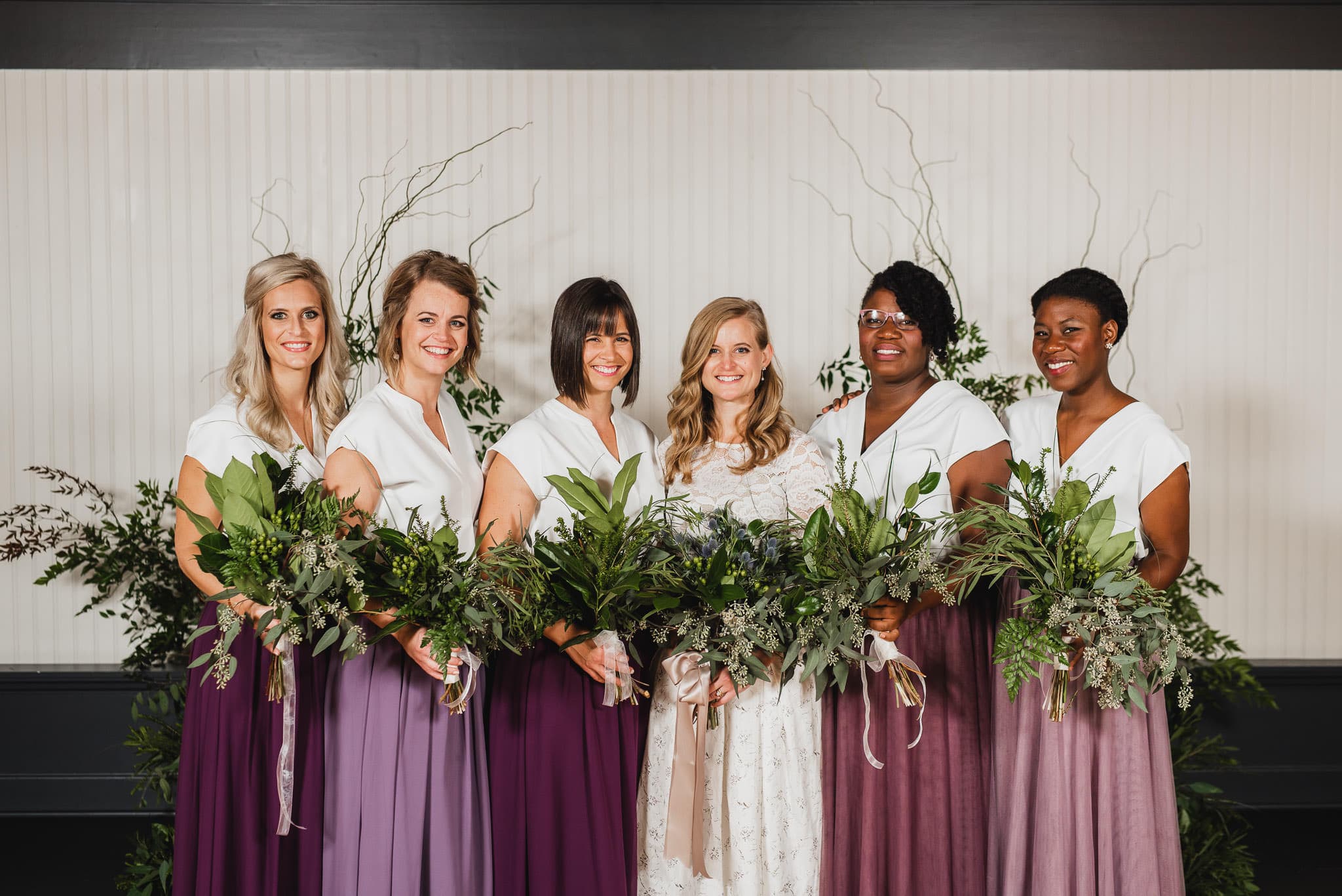 Bride in a white dress surrounded by five bridesmaids in purple skirts, all holding bouquets, posed elegantly against a floral backdrop at the wedding at the Evergreen.