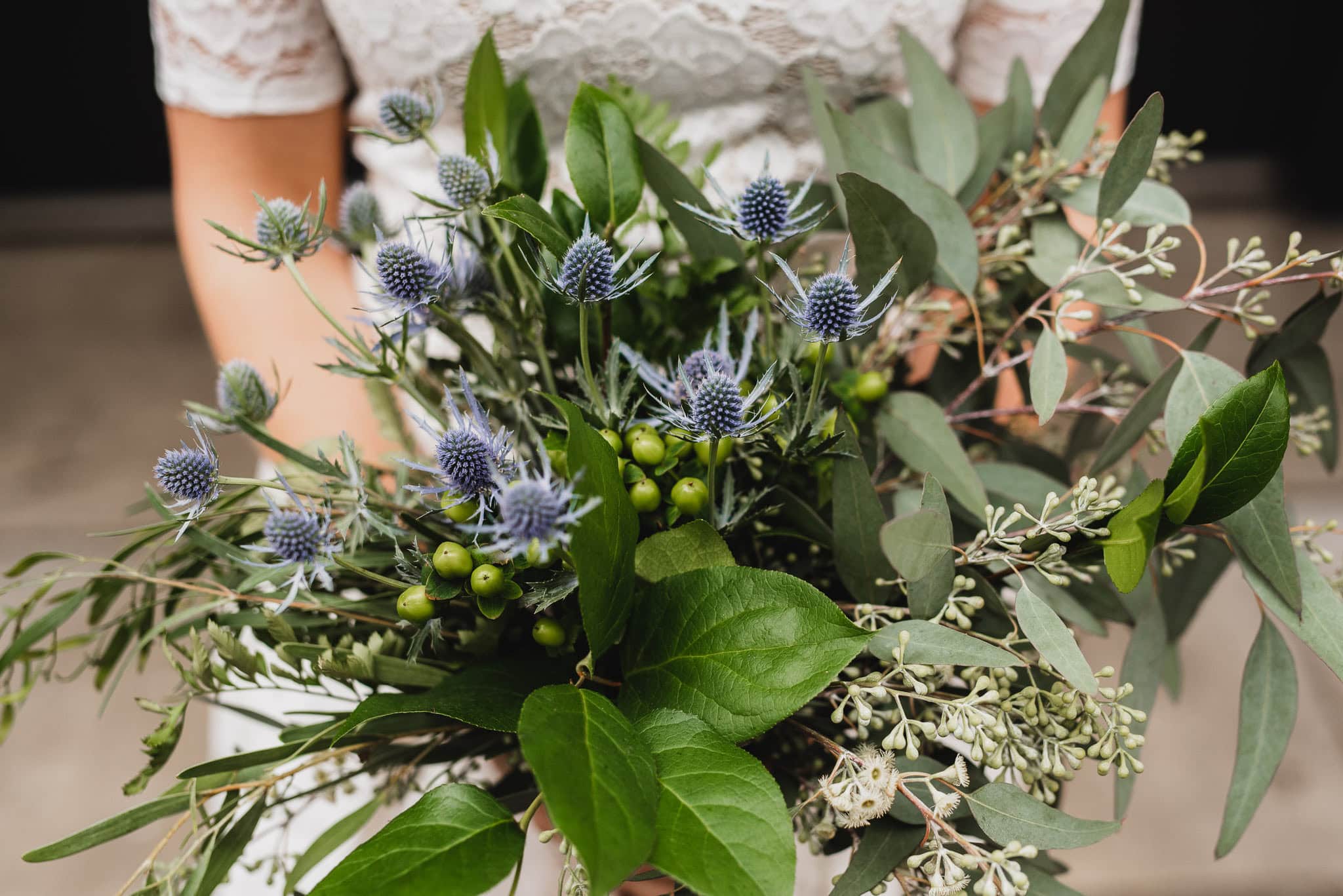 Close-up of a person holding a leafy bouquet with green leaves, small green berries, and spiky blue flowers, perfect for wedding portraits near The Evergreen.