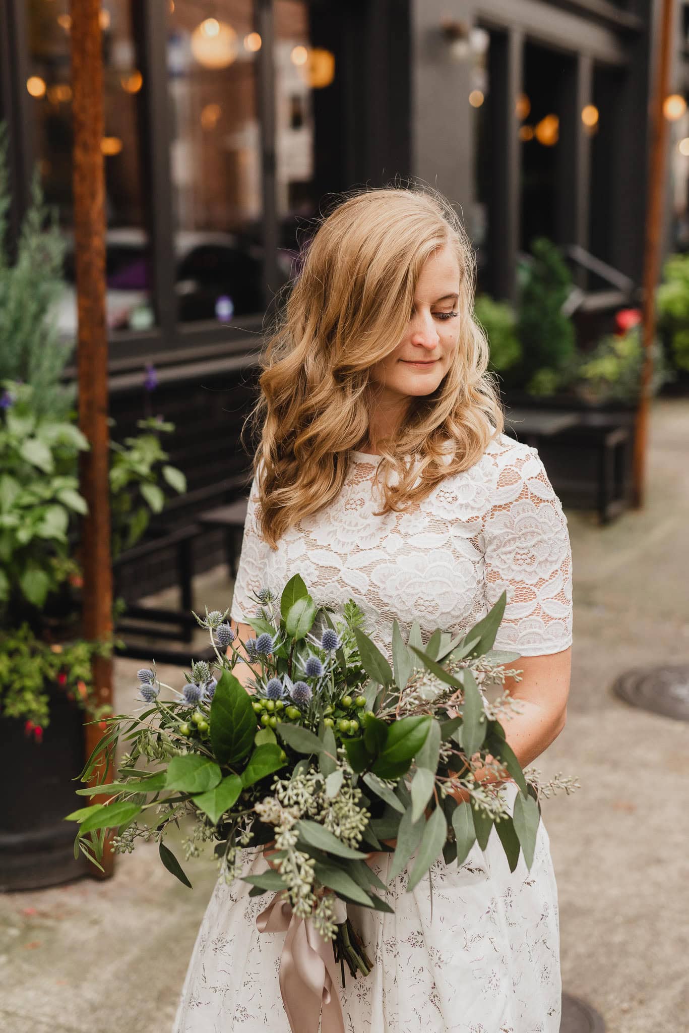 A woman in a white lace dress stands on a city sidewalk, holding a bouquet of greenery and blue flowers—perfect for wedding portraits near The Evergreen.