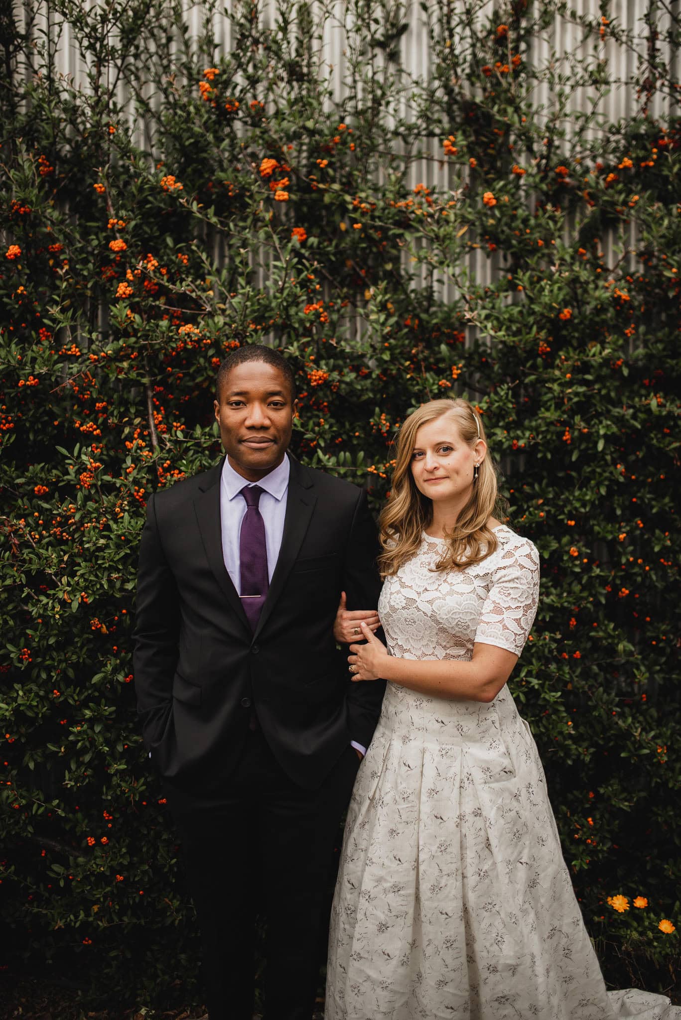 A couple poses in front of a flowered hedge near The Evergreen; the man is in a suit and the woman is in a white patterned dress, perfect for their wedding portraits.