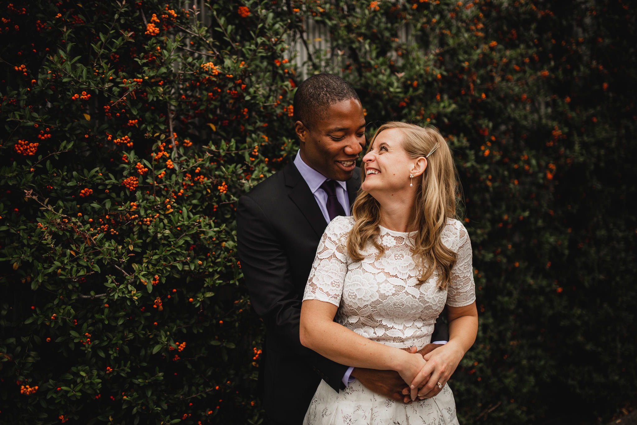 The happy couple embraces in front of a lush wall adorned with greenery and vibrant orange berries, making it a perfect spot for wedding portraits near The Evergreen.
