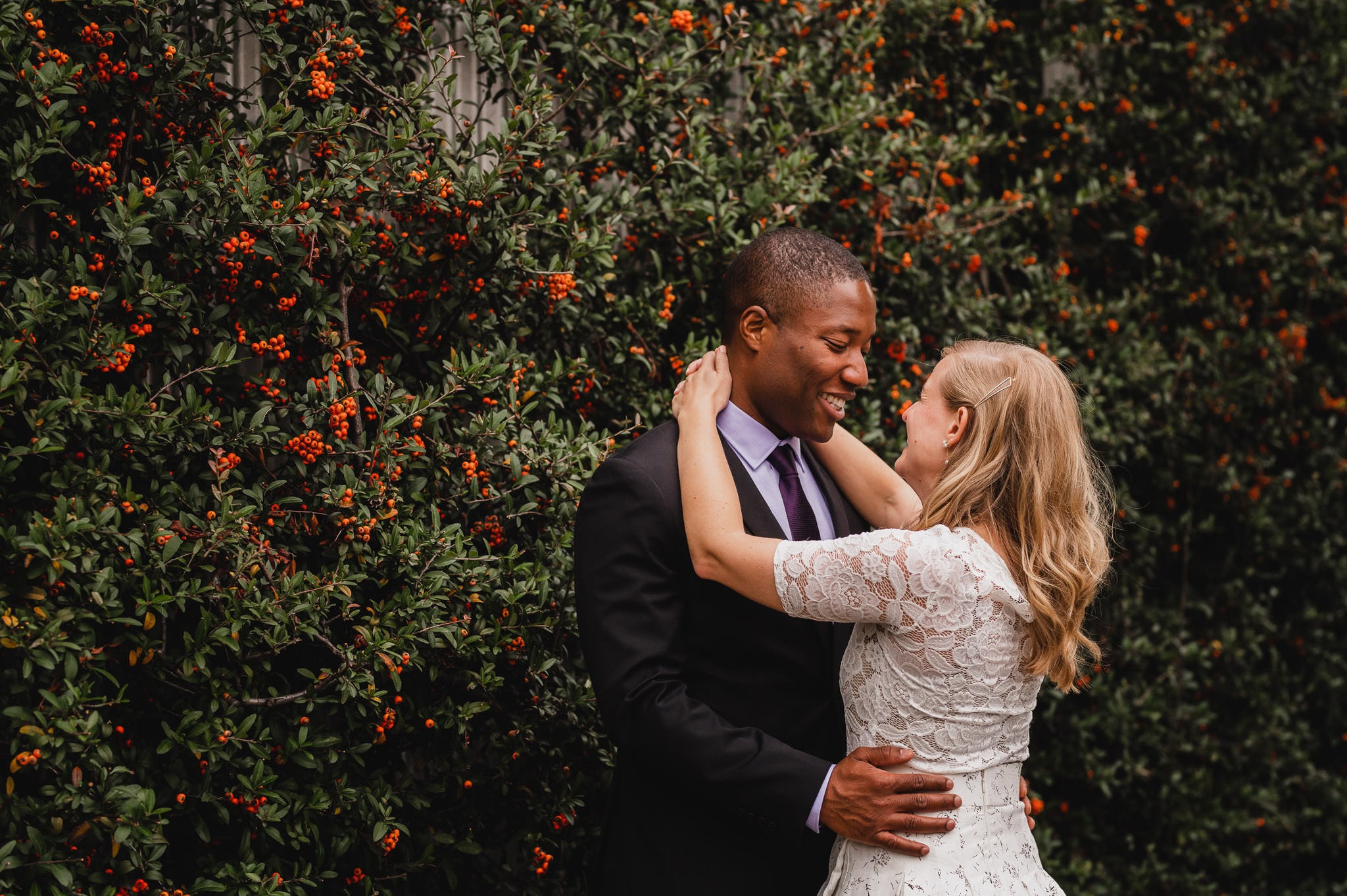 A couple embraces and smiles in front of a lush, berry-covered bush, capturing a perfect moment for their wedding portraits near The Evergreen.