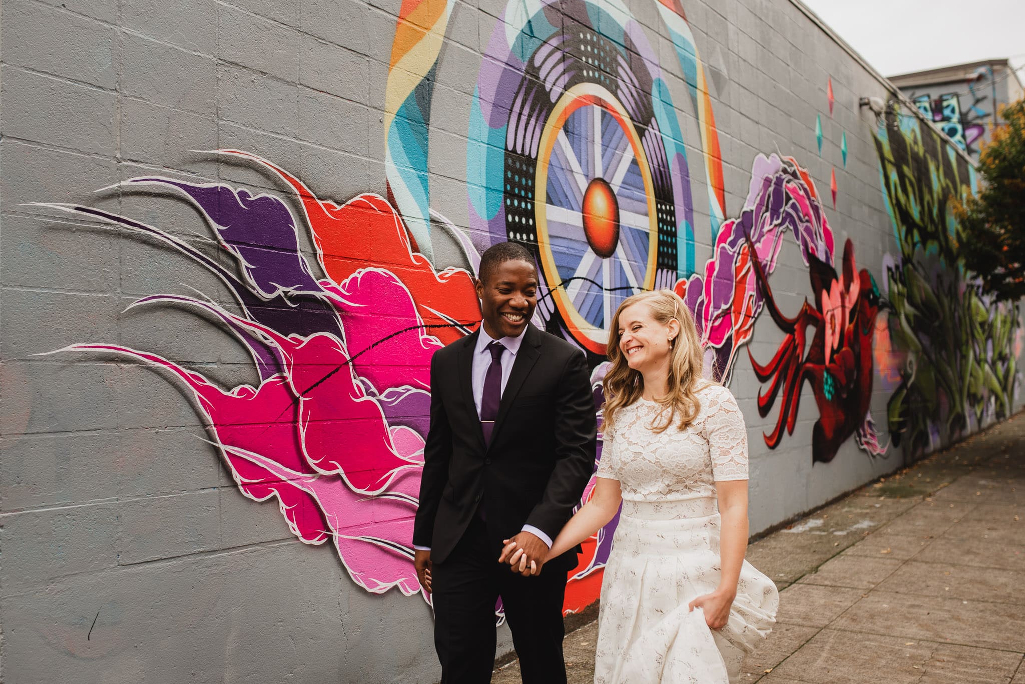 A couple in formal attire holds hands, smiling in front of a colorful graffiti mural on a city street, perfect for capturing wedding portraits near The Evergreen.