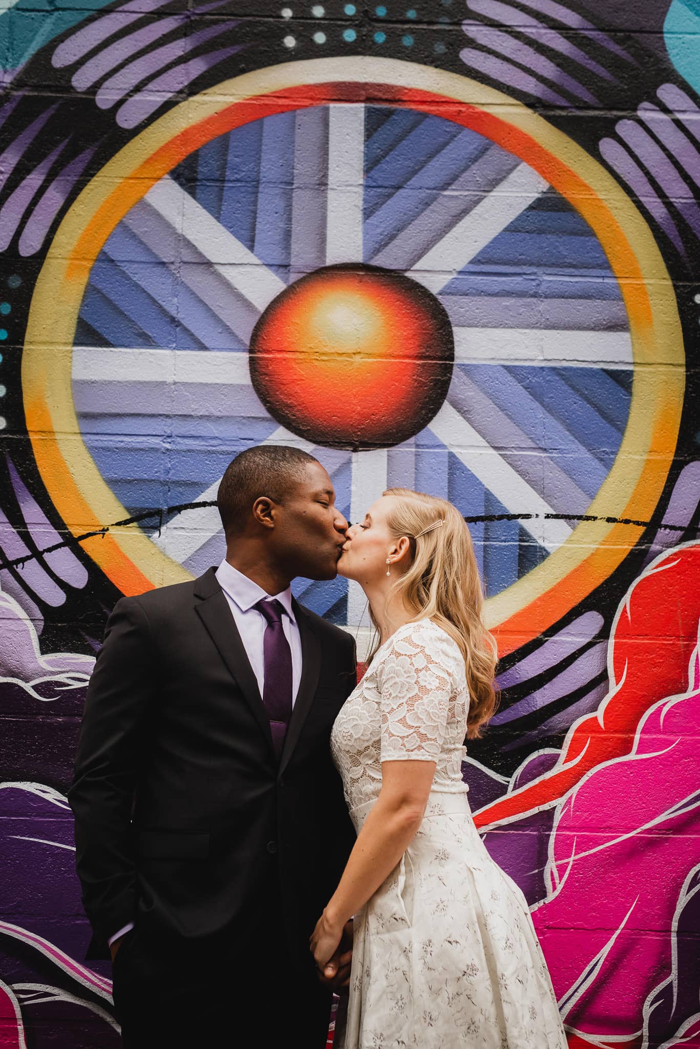 A couple kisses in front of a colorful mural with circular and geometric patterns, making it a perfect spot for wedding portraits near The Evergreen.
