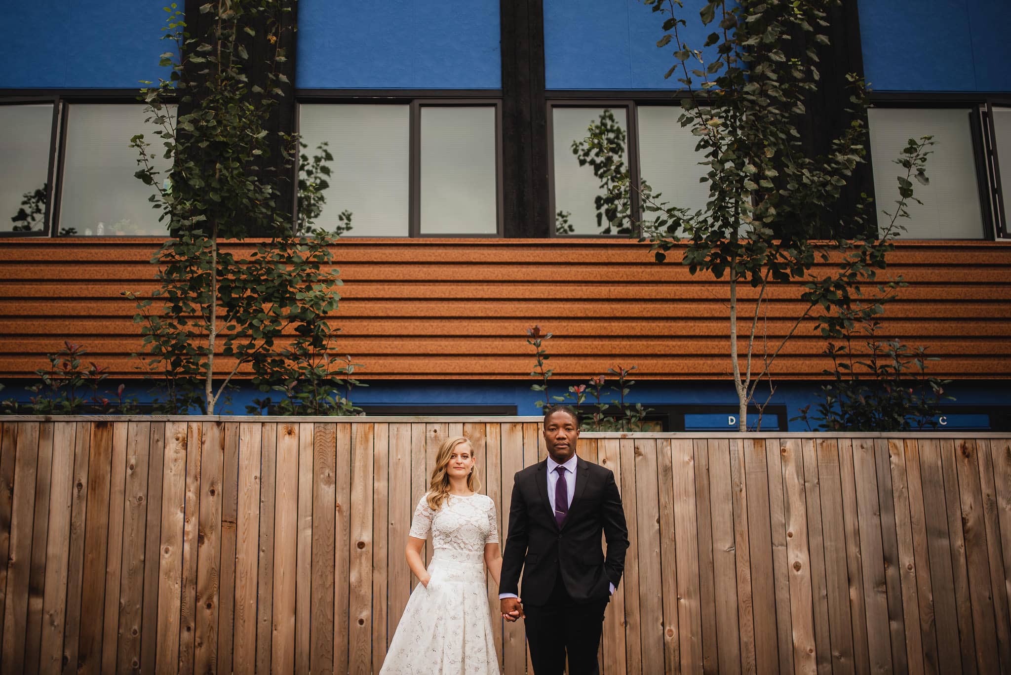 A bride and groom stand hand in hand in front of a wood fence and modern building, capturing their wedding portraits near The Evergreen.