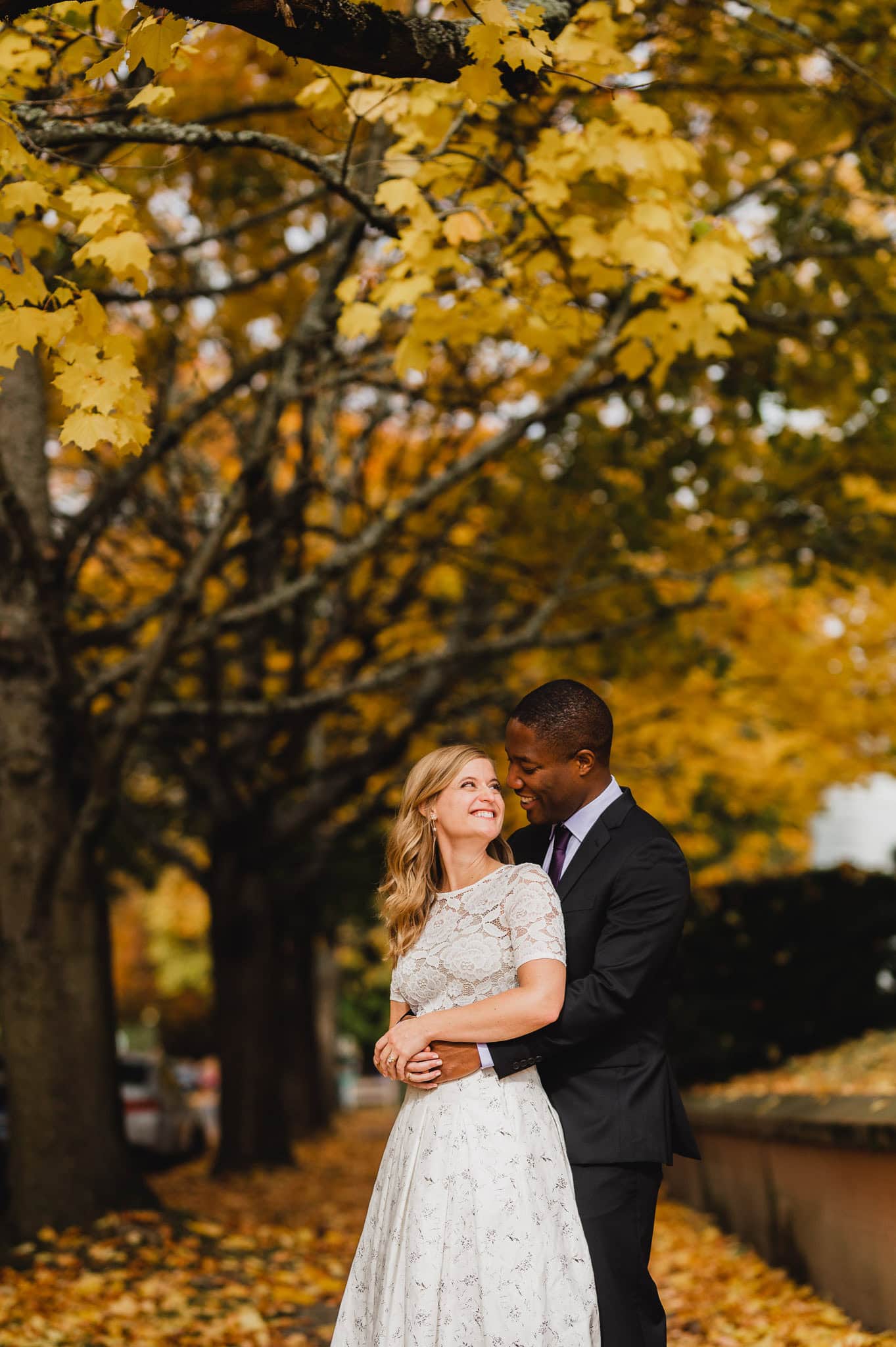 A couple embraces under autumn trees with yellow leaves, both smiling and enjoying the fall scenery, perfect for wedding portraits near The Evergreen.