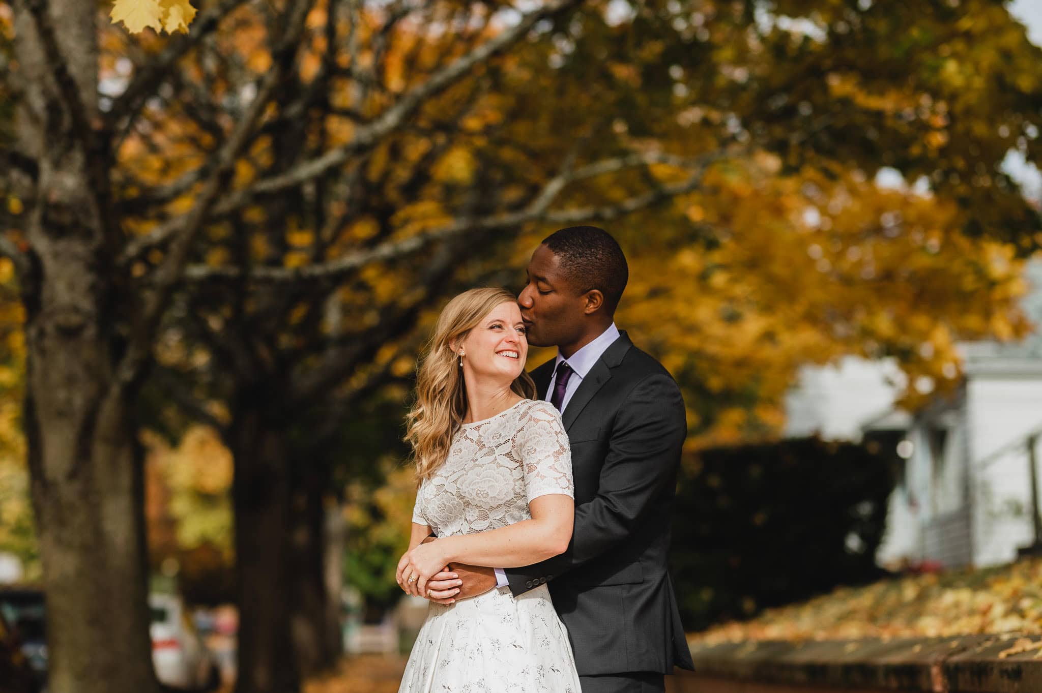 A couple poses for wedding portraits near The Evergreen, with a man embracing a woman from behind under autumn trees; both are smiling. She wears a white dress while he dons a dark suit.