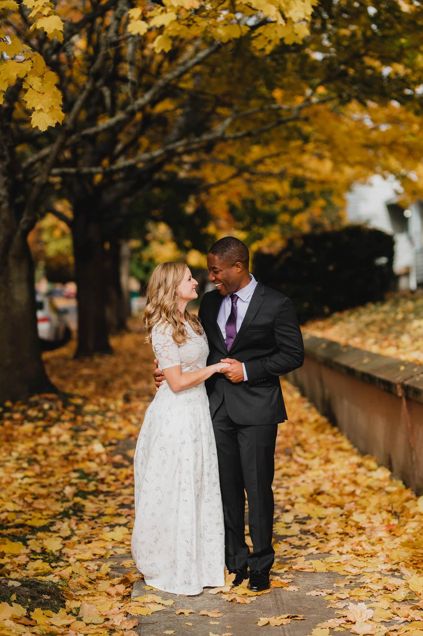 A couple in formal attire stands on a leaf-covered path surrounded by autumn trees near The Evergreen, smiling warmly at each other.