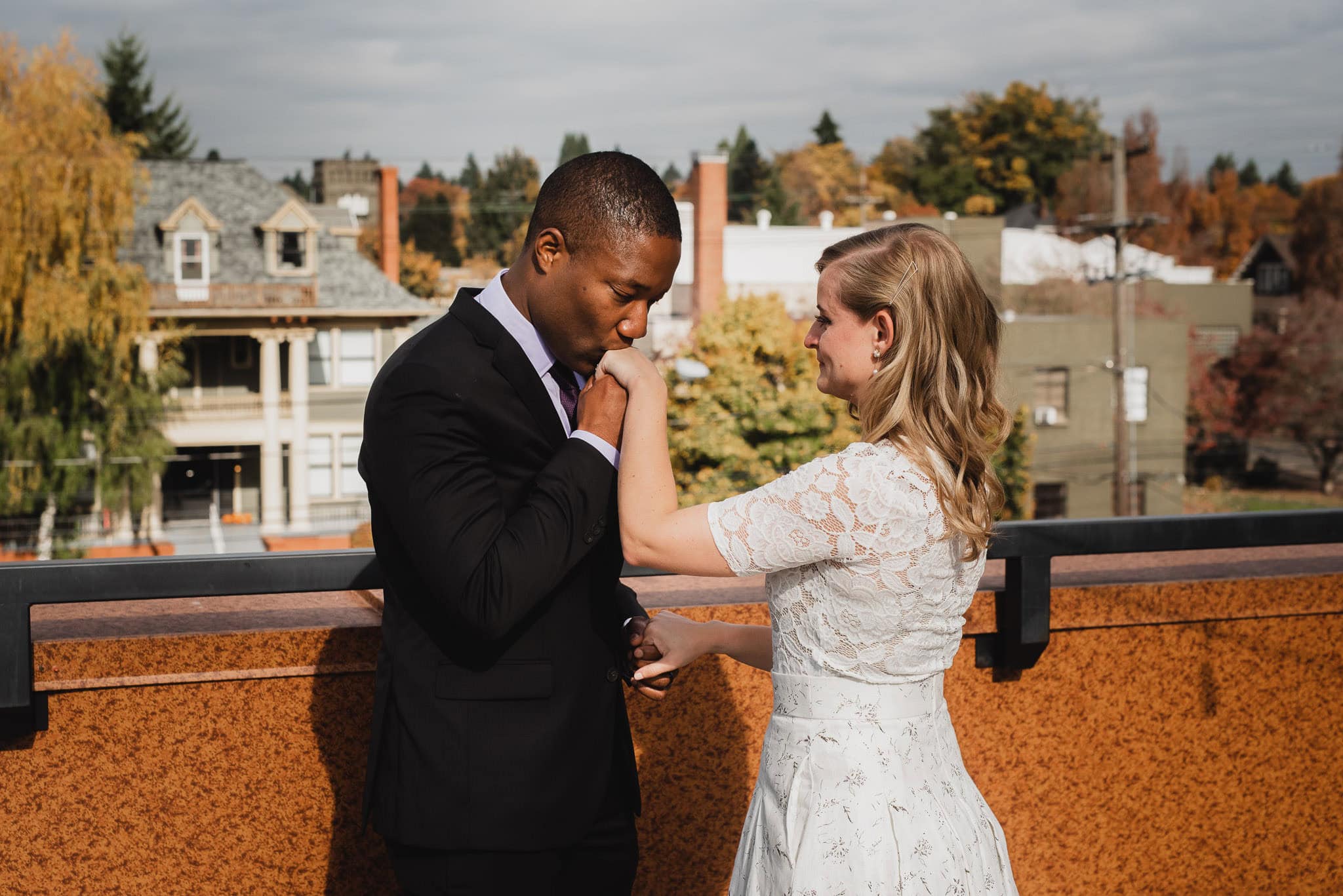 A couple shares a romantic first look on the rooftop of an Airbnb before their wedding at The Evergreen; the man in a suit tenderly kisses the womans hand as she stands elegantly in her white floral dress.