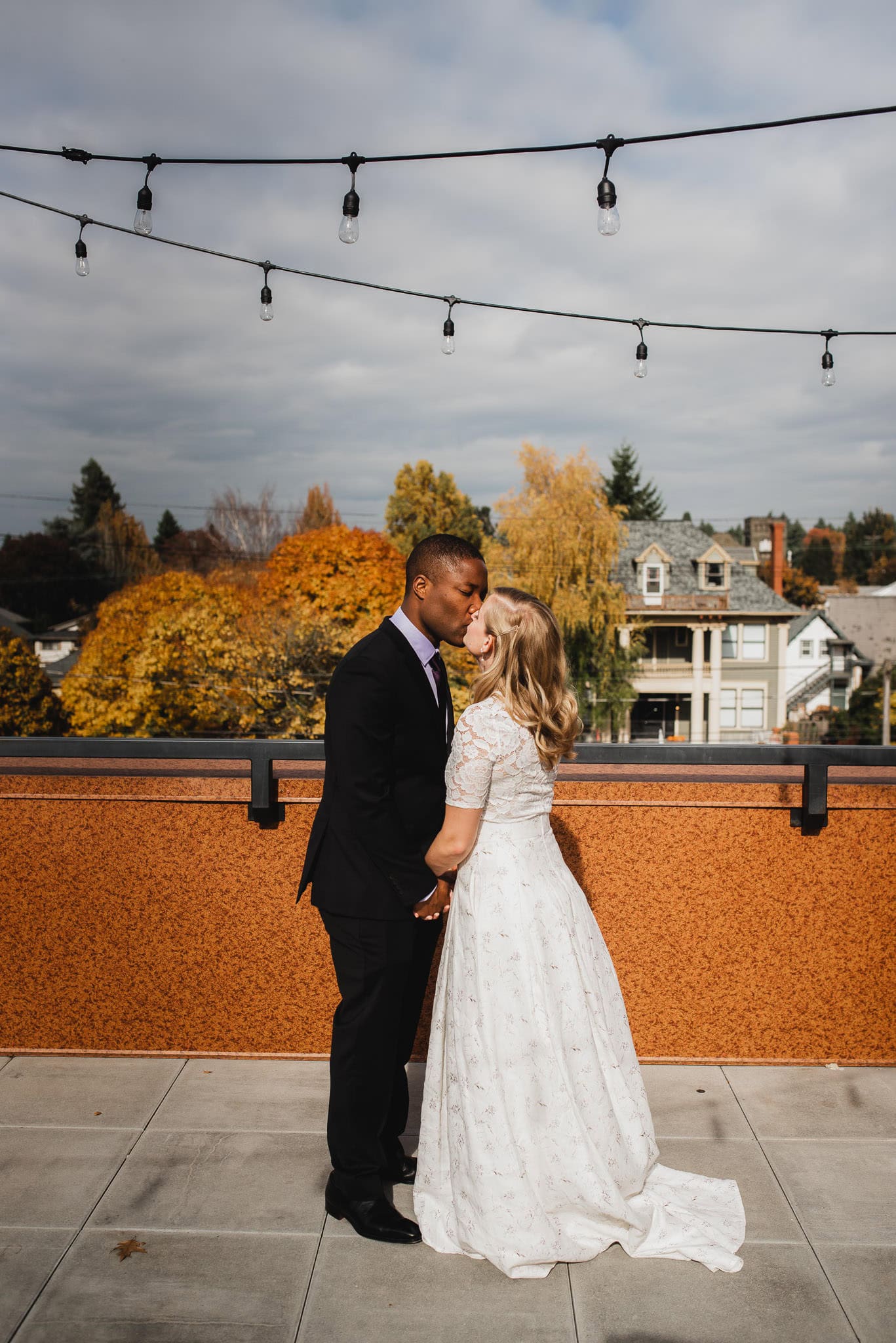 A couple in formal attire shares a kiss on the rooftop of an Airbnb, surrounded by autumn trees and twinkling string lights, during their first look before a romantic wedding at The Evergreen.