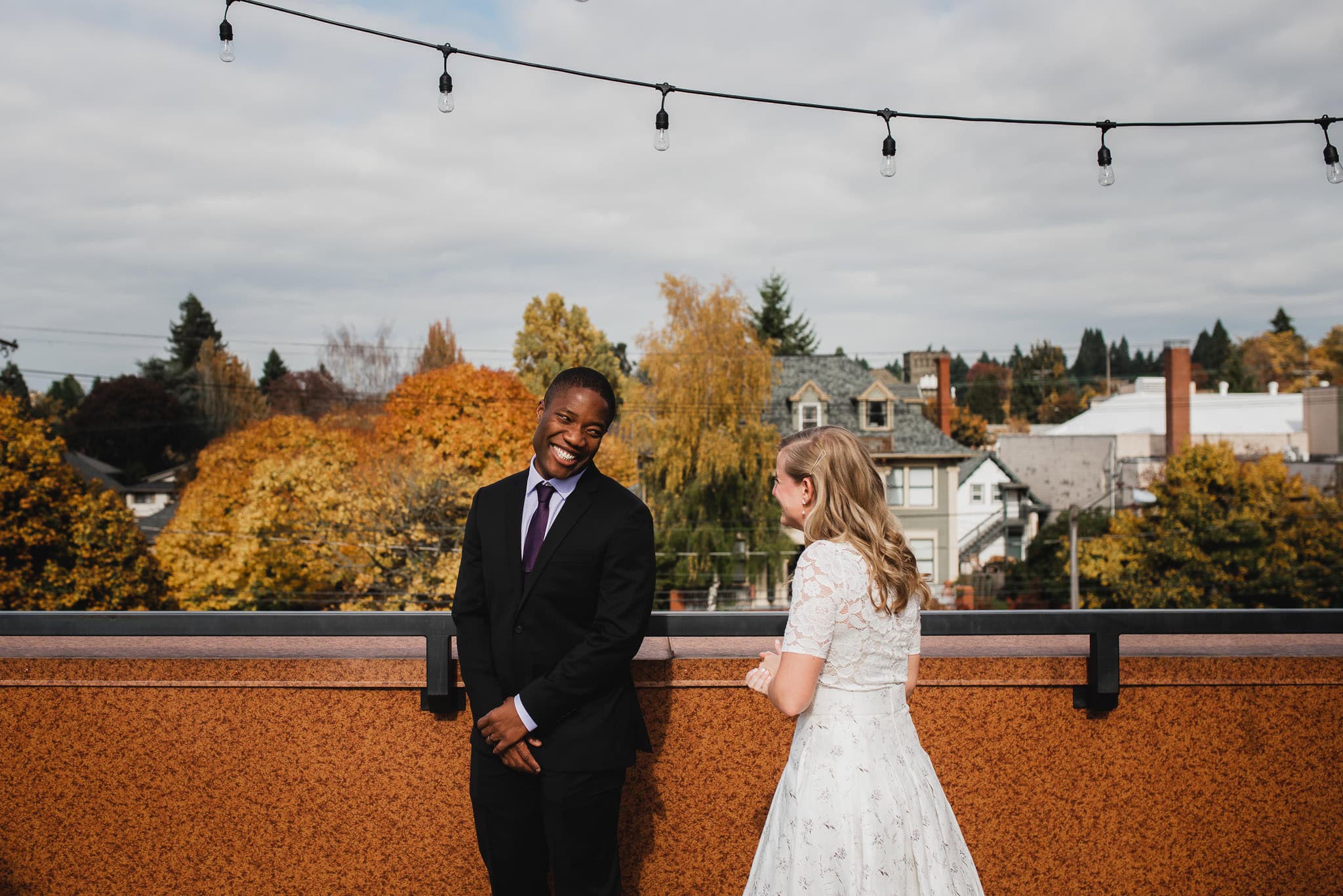 A man in a suit and a woman in a white dress share their first look on the rooftop of an Airbnb, smiles bright against the backdrop of autumn trees, as they prepare for their wedding at The Evergreen.