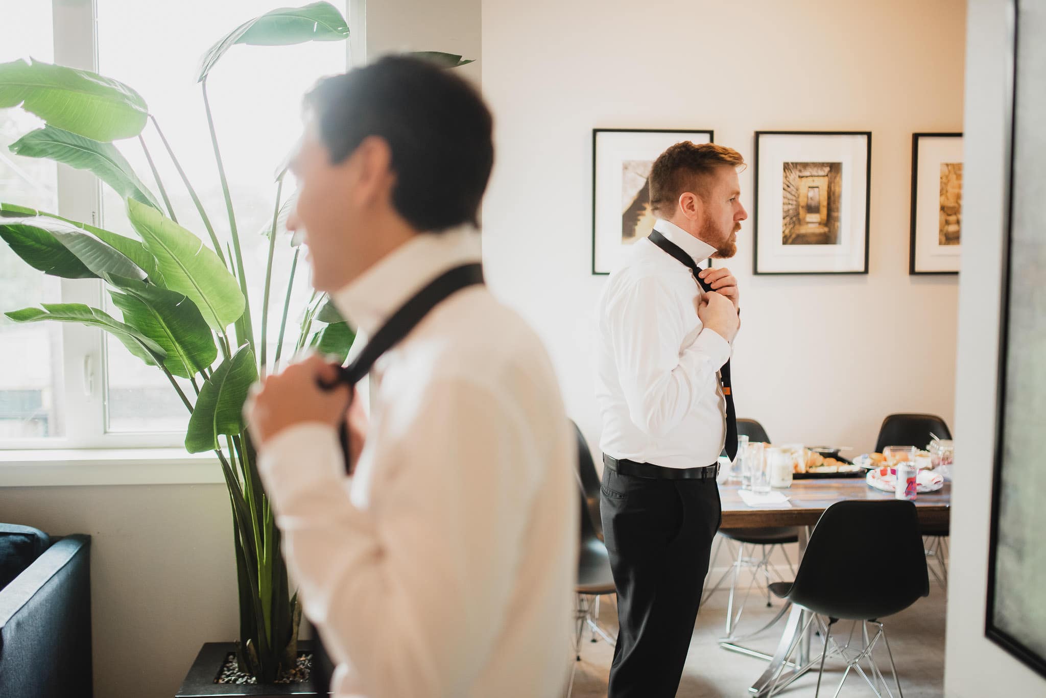 Two men in white shirts adjust their ties in a stylish room at an Airbnb rental. The space, adorned with a large plant and framed art on the wall, is buzzing with anticipation as they get ready for a wedding at The Evergreen.