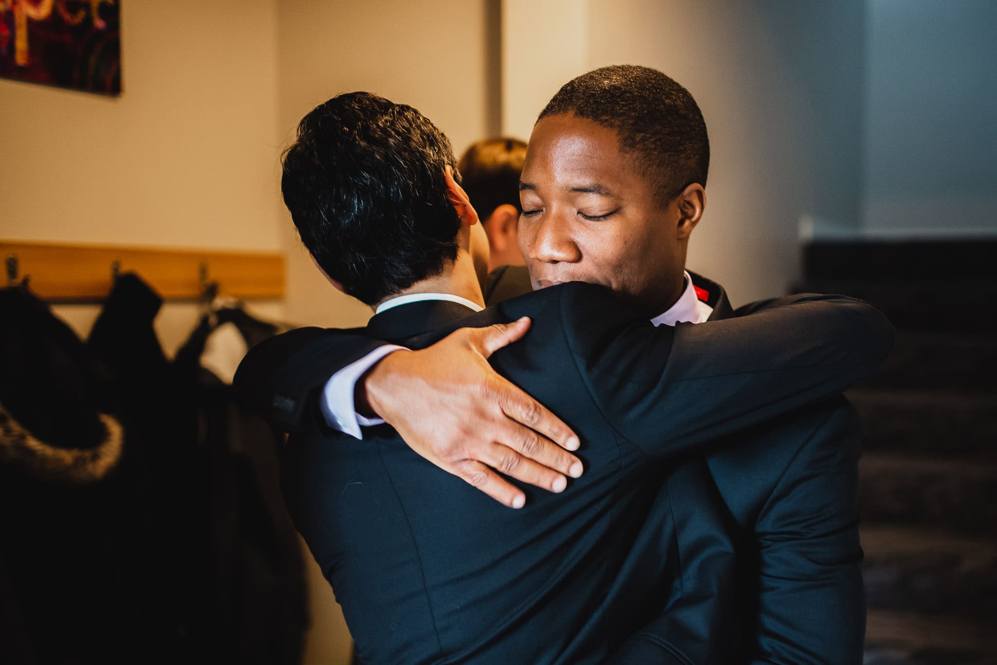Two men in suits warmly embrace in a hallway at their Airbnb rental, conveying a sense of friendship and reunion as they prepare for a wedding at The Evergreen.