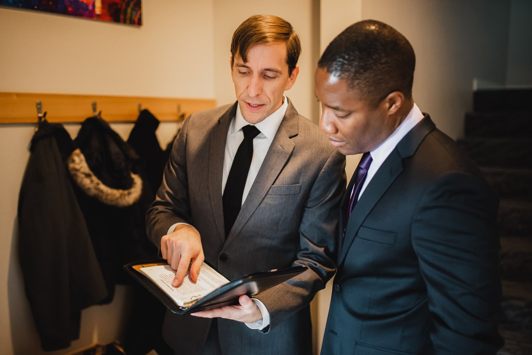 Two men in suits are reviewing a document together near a coat rack, at an Airbnb rental while getting ready for a wedding at The Evergreen.