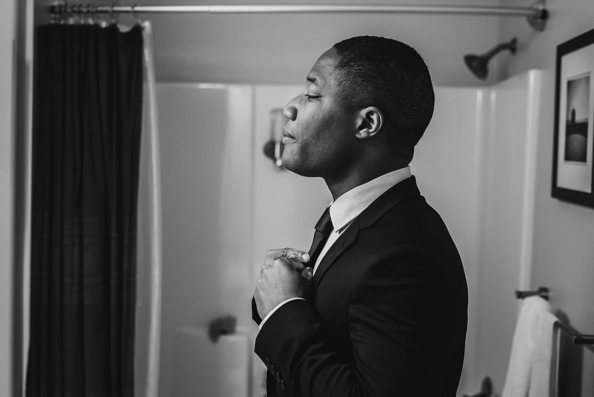 Man in a suit adjusts his tie in the bathroom at an Airbnb rental, reflected gracefully in the mirror. Black and white photo captures him getting ready for a wedding at The Evergreen.