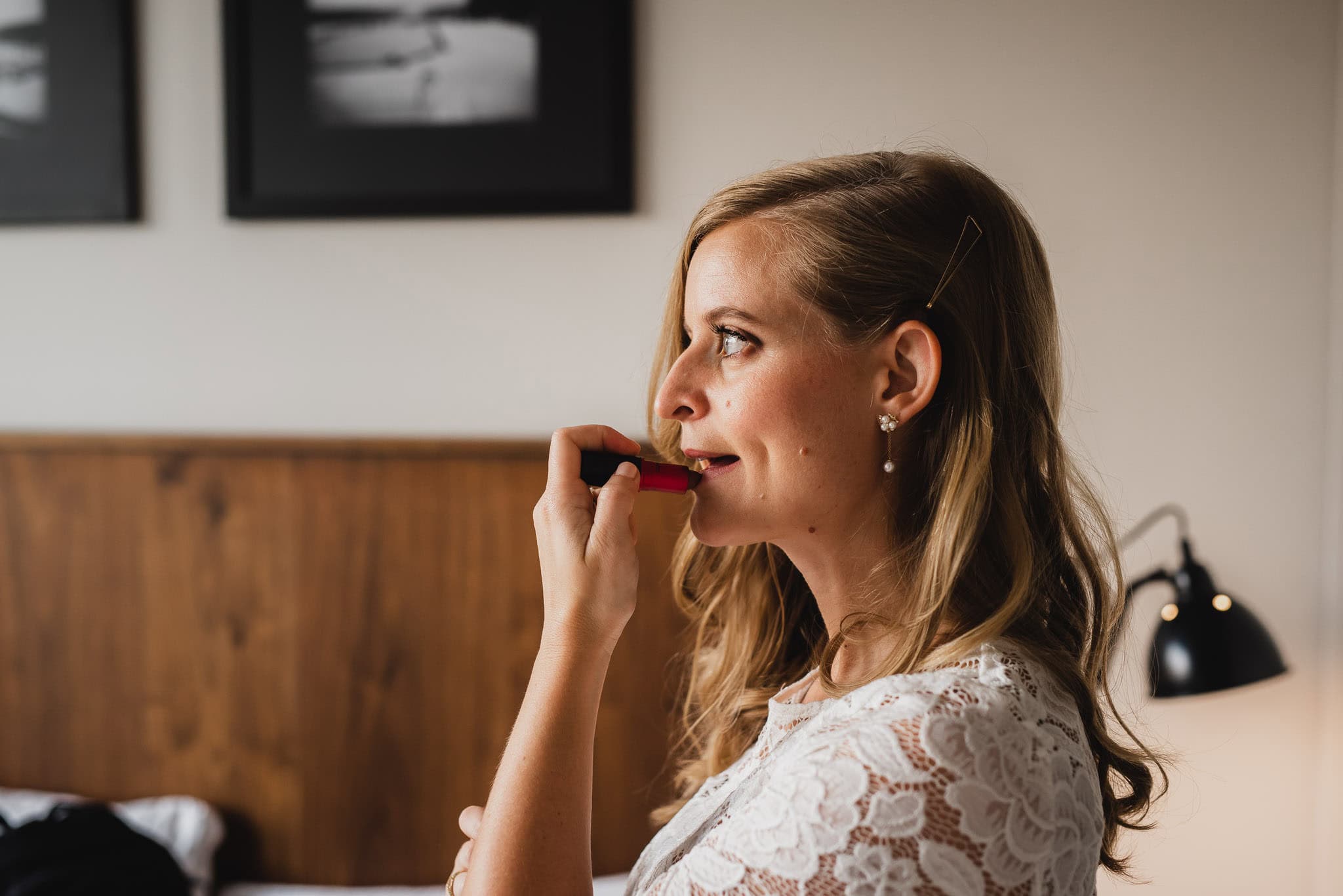 In a beautifully decorated Airbnb rental, a woman in a white lace dress applies lipstick, framed photos and a glowing lamp behind her. Shes getting ready for a wedding at The Evergreen, capturing the elegance of the moment.