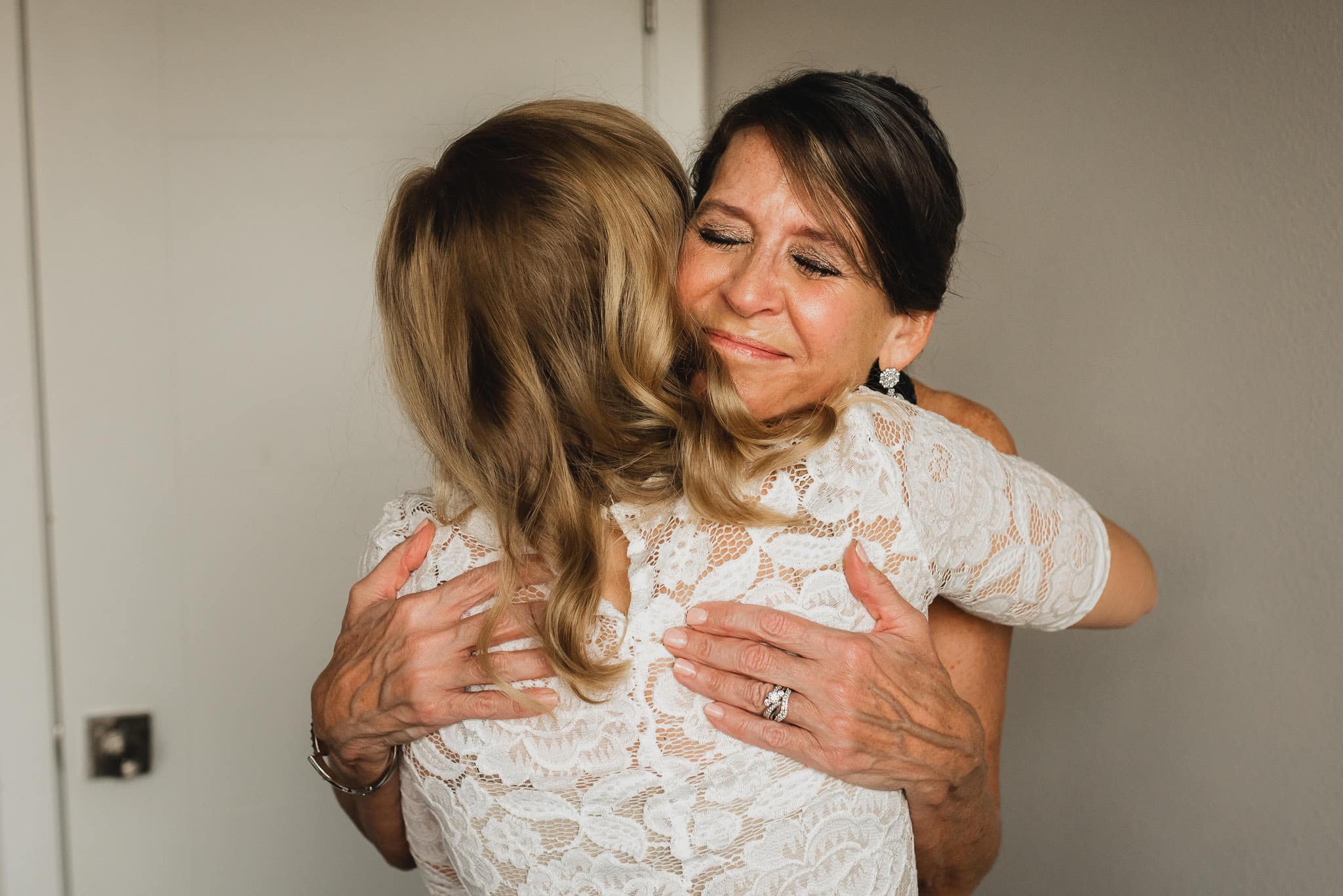 Two women embrace warmly, one with closed eyes, in white lace outfits at an Airbnb rental, getting ready for a wedding at The Evergreen.
