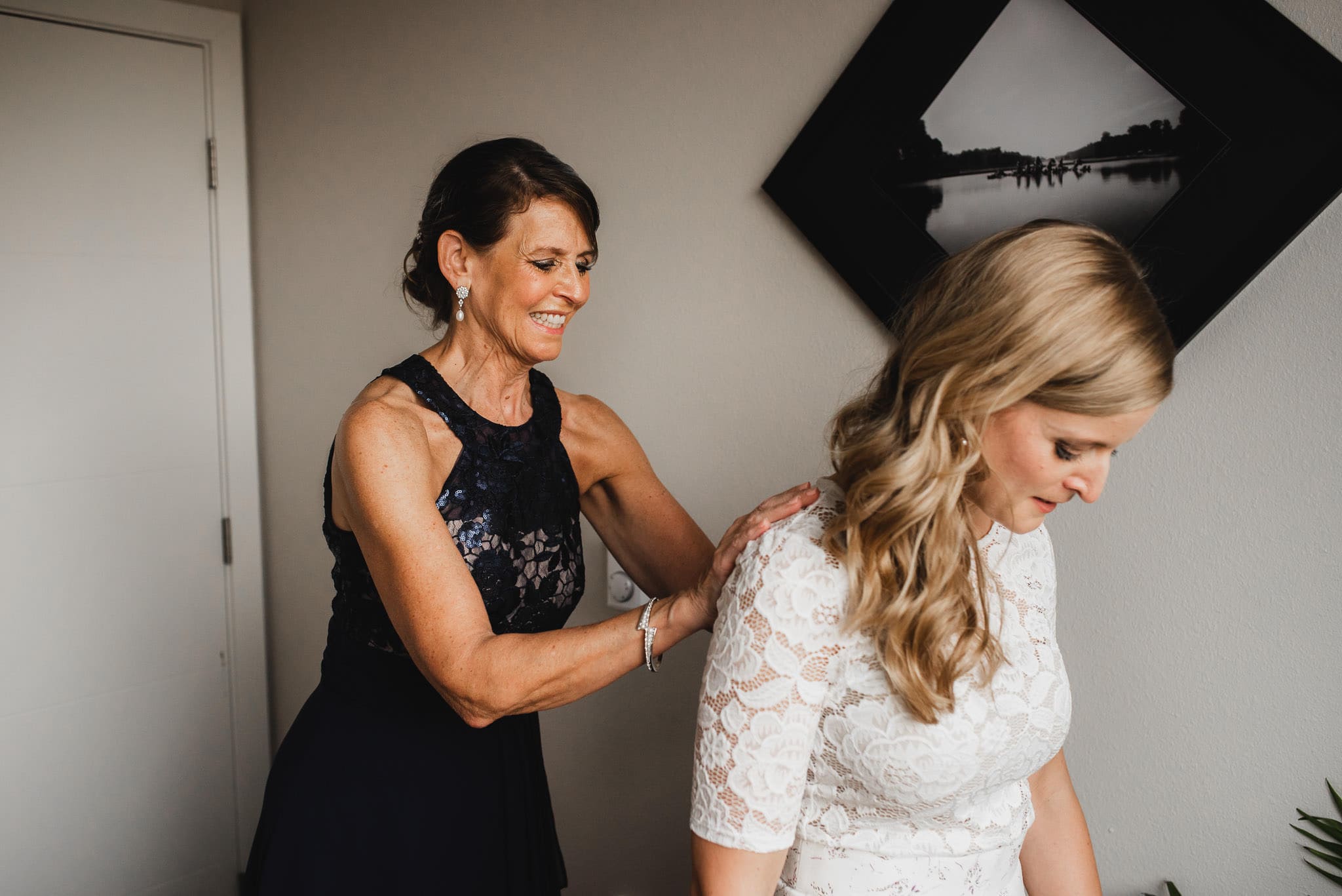 In a cozy room at an Airbnb rental, a woman carefully assists a bride with her dress as they prepare for the wedding at The Evergreen. A framed photo on the wall adds to the serene ambiance of anticipation and joy.