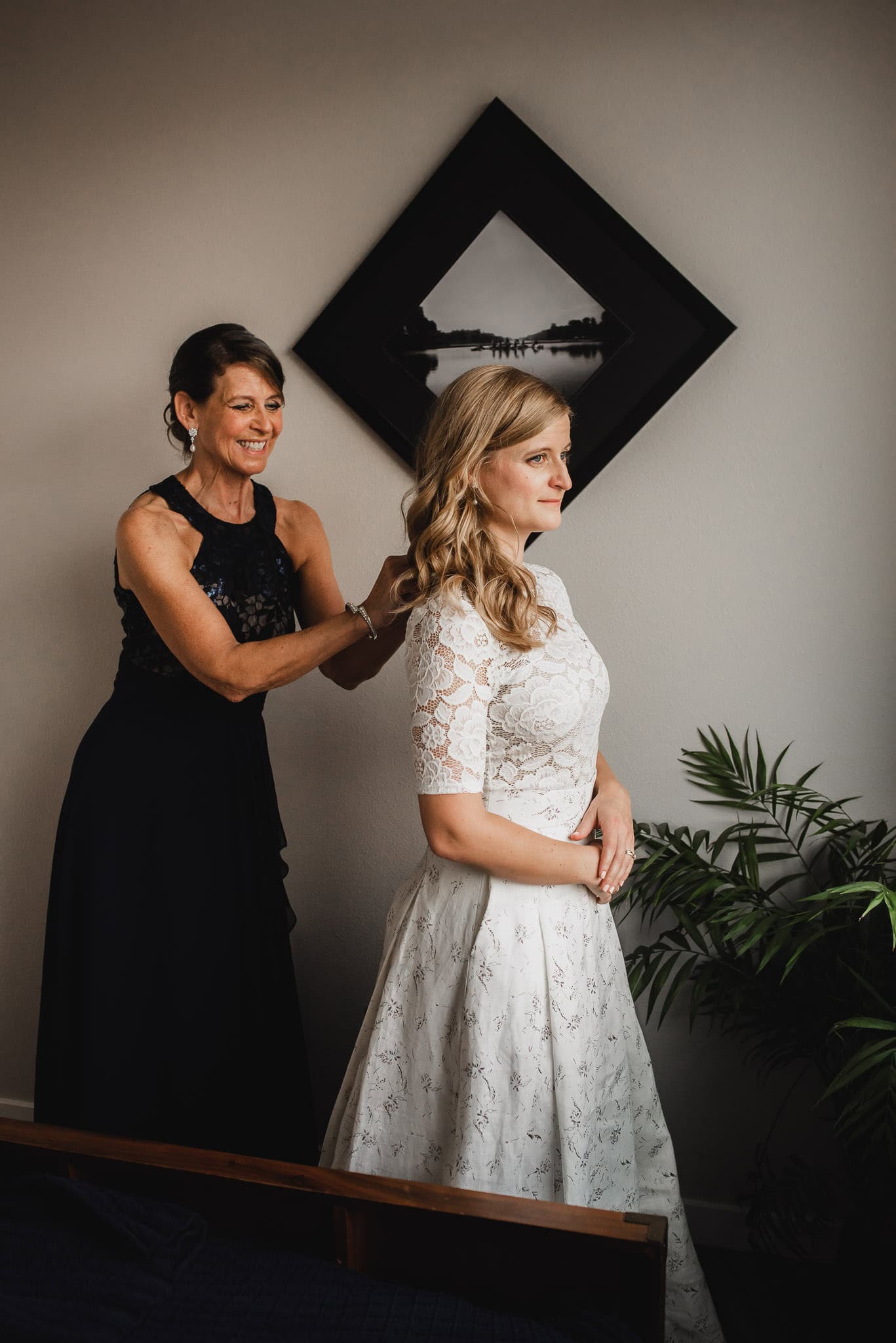 In an Airbnb rental, a woman in a black dress assists the bride with her hair beneath framed art and a lush plant, preparing for the wedding at The Evergreen.
