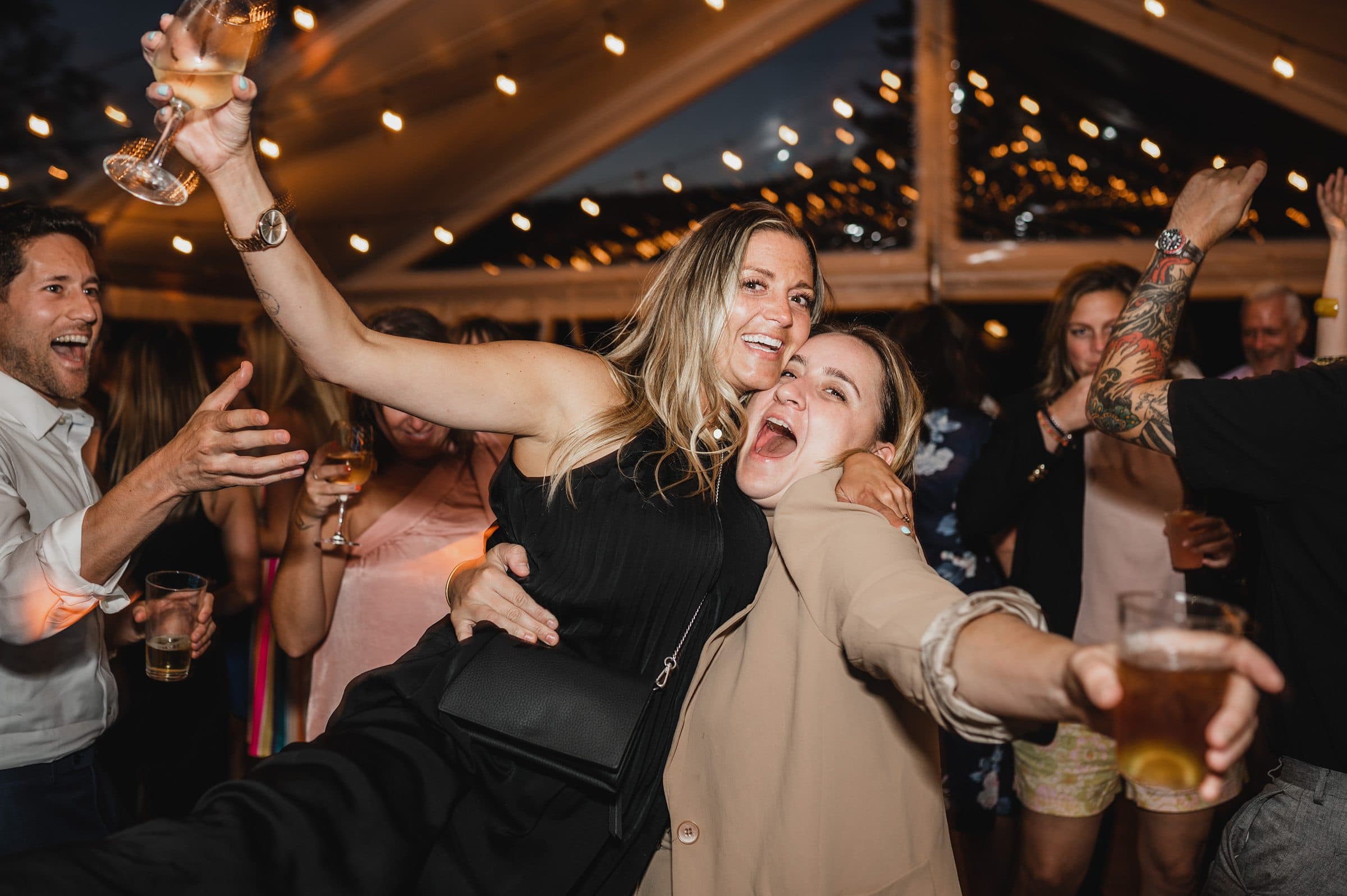 Two women joyfully posing and dancing at a lively party under string lights, surrounded by people, capturing the spirit of wedding photos at The Griffin House.