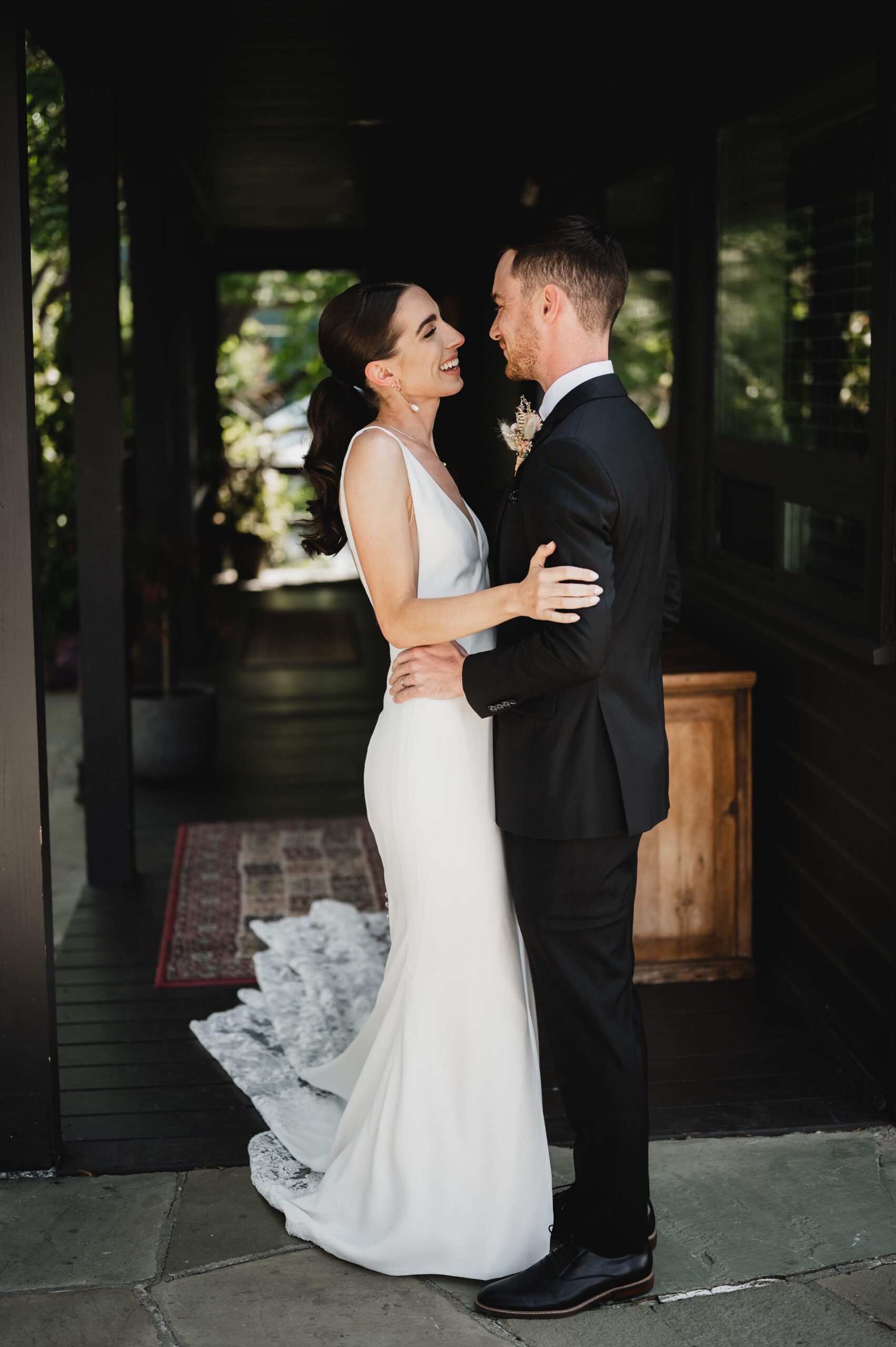 A bride and groom smile at each other, embracing in a dimly lit corridor with greenery in the background, capturing the magic of wedding photos at The Griffin House.