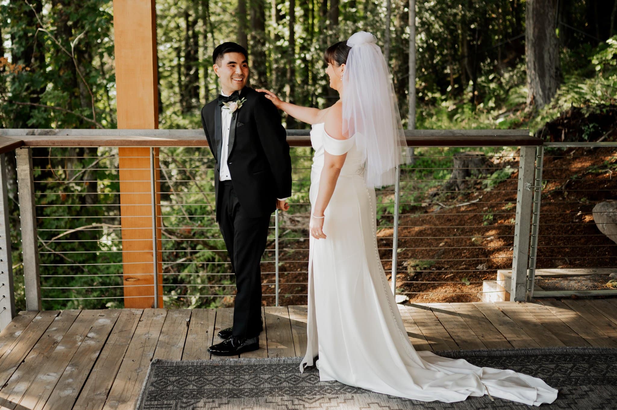 Bride in a white dress and veil taps her groom's shoulder on a wooden deck, surrounded by lush green trees, during their first look at Amaterra Winery, a charming Portland wedding venue.