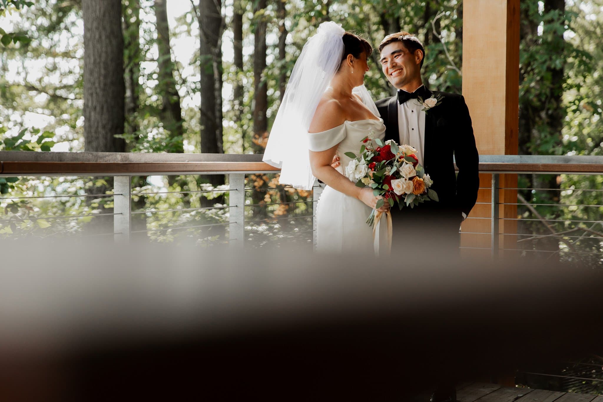 A bride and groom share a joyful smile on a forested balcony, holding a bouquet of flowers during their first look at the enchanting Amaterra Winery, a Portland wedding venue.