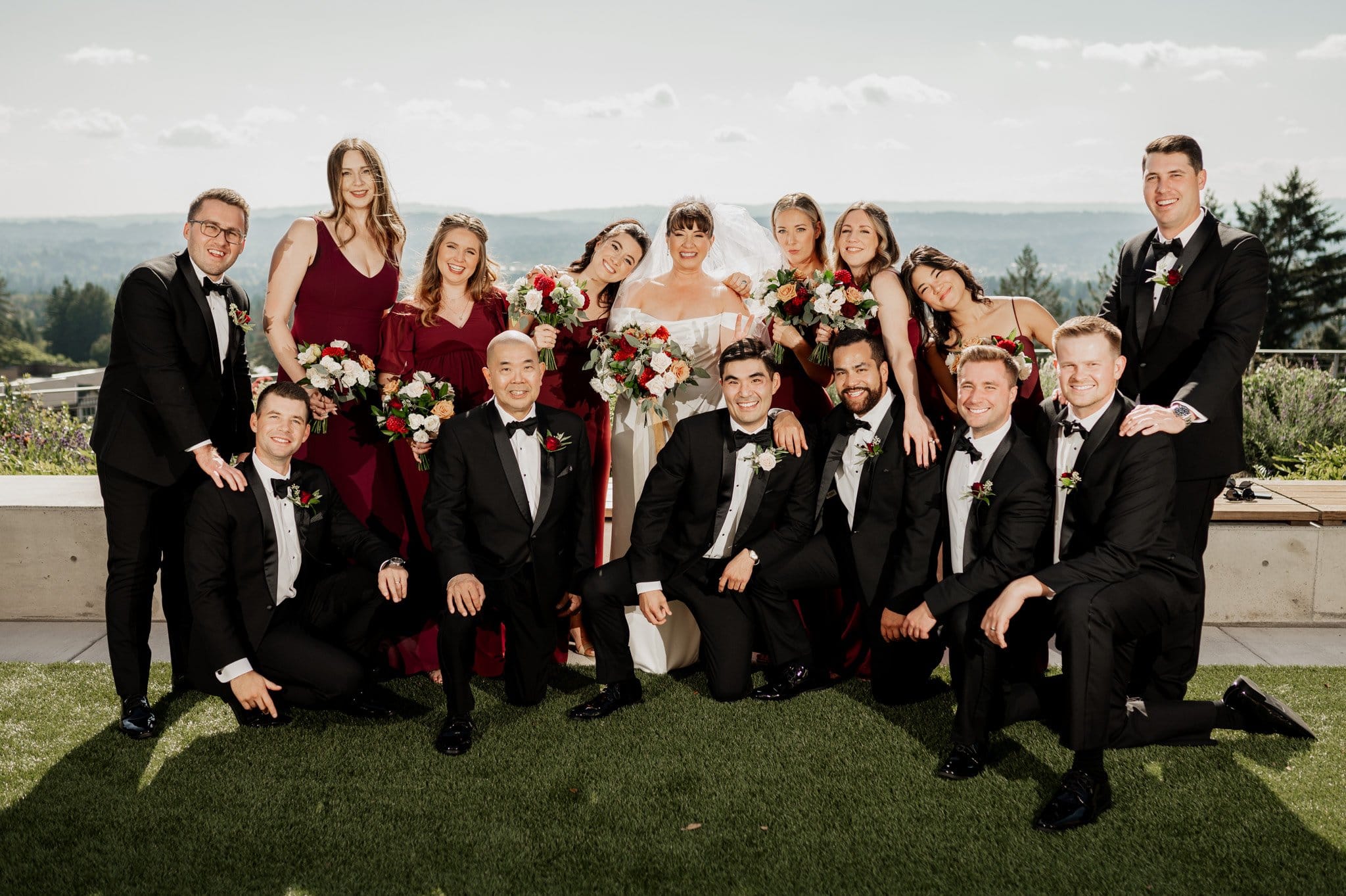 A wedding party poses outdoors at the picturesque Amaterra Winery, a renowned Portland wedding venue. Bridesmaids in vibrant red dresses, groomsmen in classic black suits, and the radiant bride in white create a stunning scene against the vineyards backdrop.