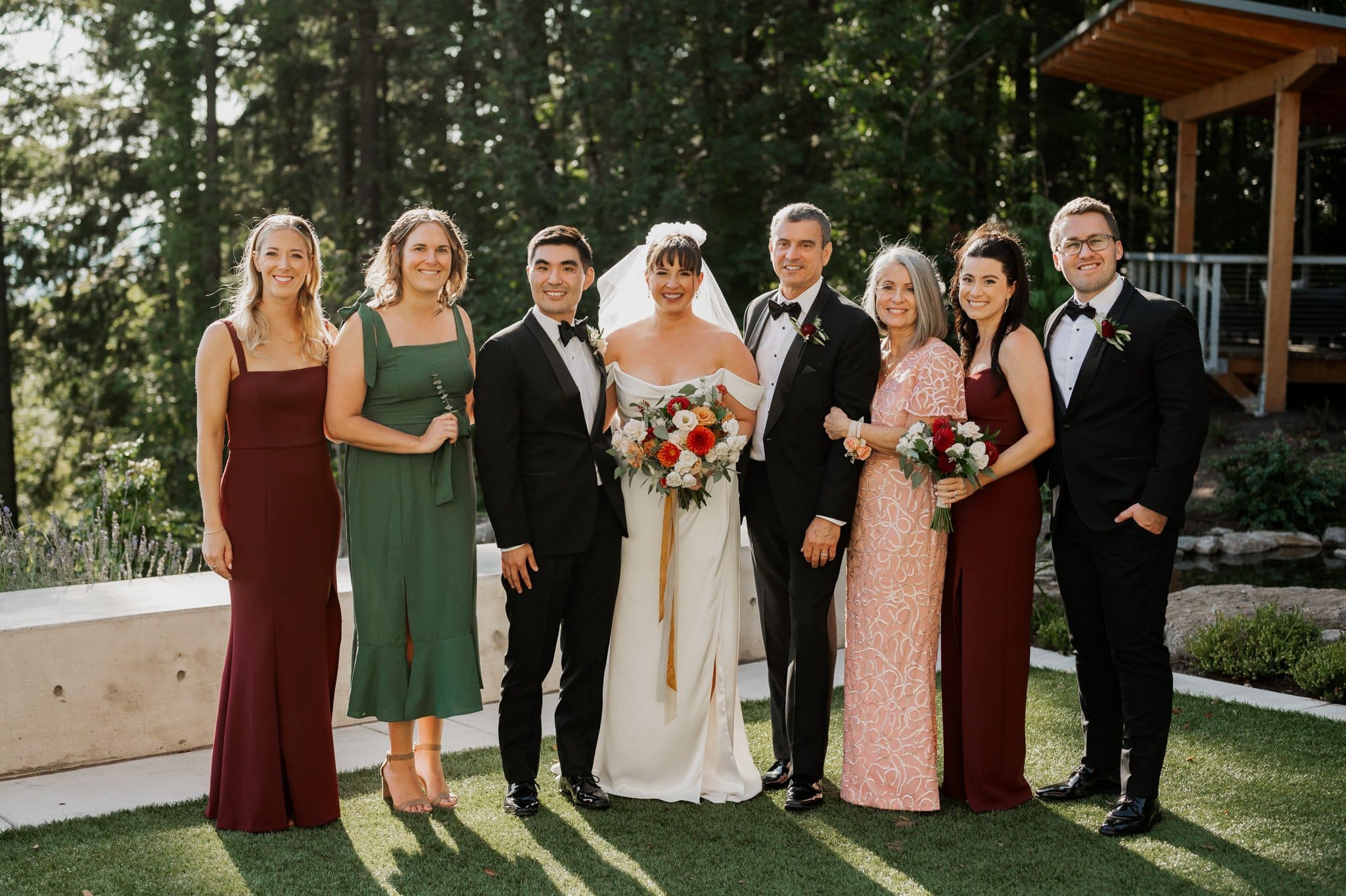 A group of eight elegantly dressed individuals poses outdoors at a wedding, with a lush forest serving as the perfect backdrop for family photos at Amaterra Winery, a renowned Portland wedding venue.
