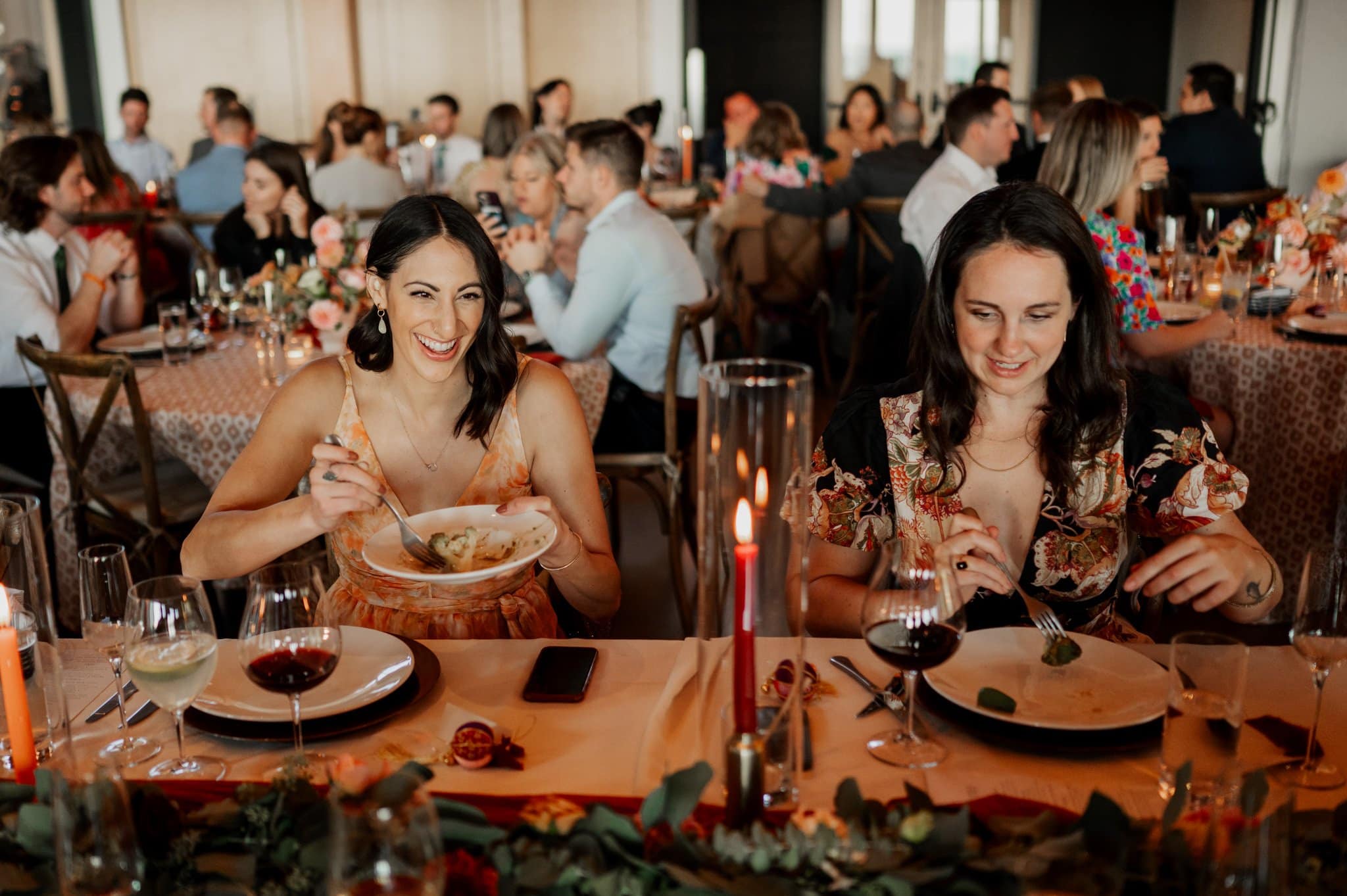 Two women savor a meal at a festive, candle-lit dinner event, set in the elegant Skyline Ballroom at Amaterra Winery, a renowned Portland wedding venue. Laughter and conversation fill the space as people socialize at tables in the warmly lit background.