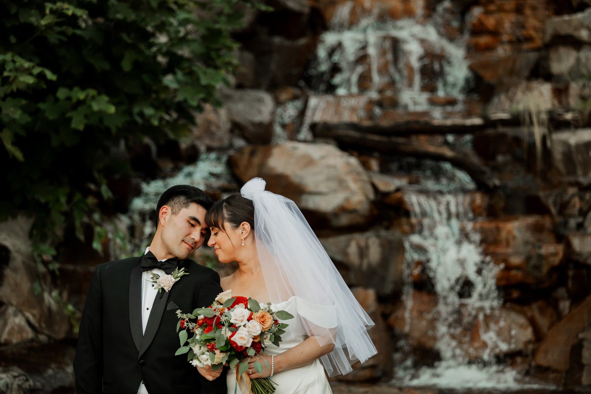 A bride and groom share a tender moment in front of a waterfall at Amaterra Winery, with the bride holding a bouquet of flowers—a picturesque scene at this Portland wedding venue.