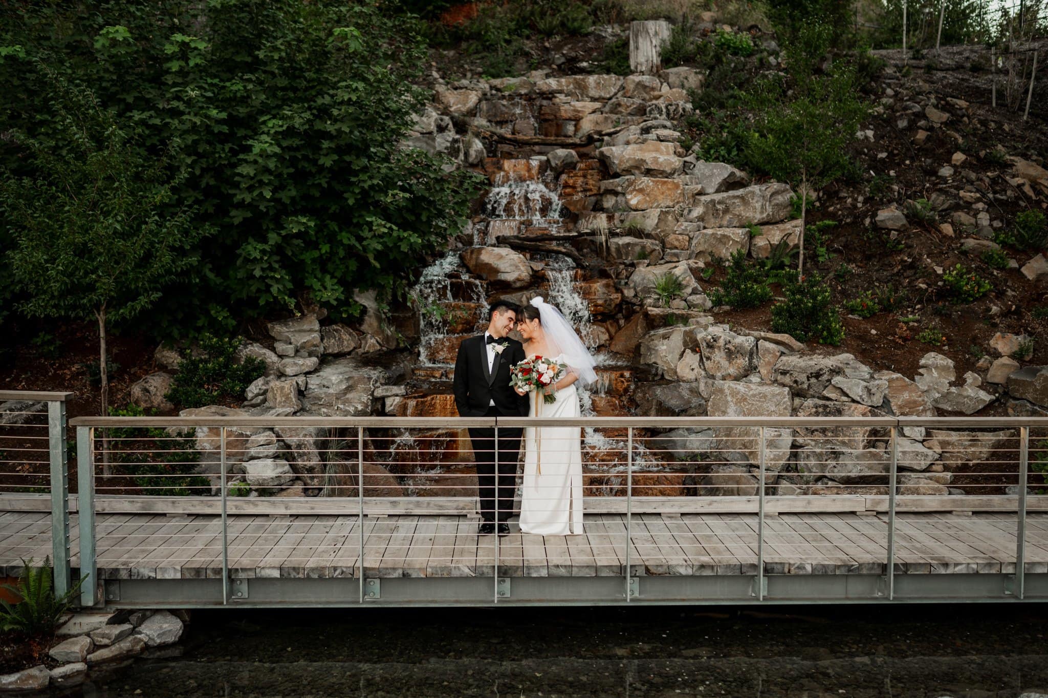 The bride and groom embrace on a bridge in front of a waterfall, surrounded by greenery and rocks, capturing the romance of Amaterra Winery, a premier Portland wedding venue.