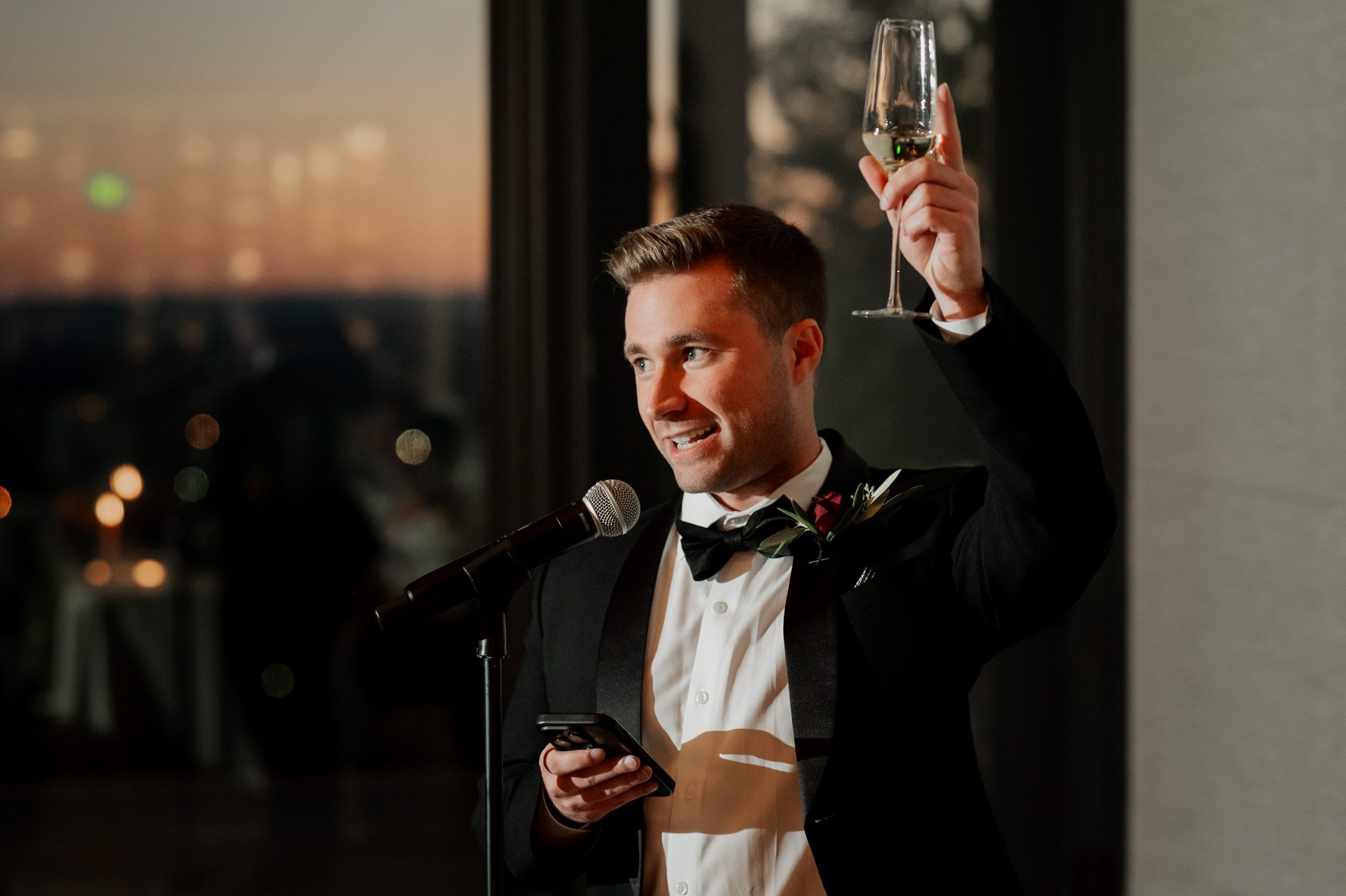 At the elegant Skyline Ballroom at Amaterra Winery, a distinguished Portland wedding venue, a person in a tuxedo raises a champagne glass while speaking into a microphone at an indoor event.