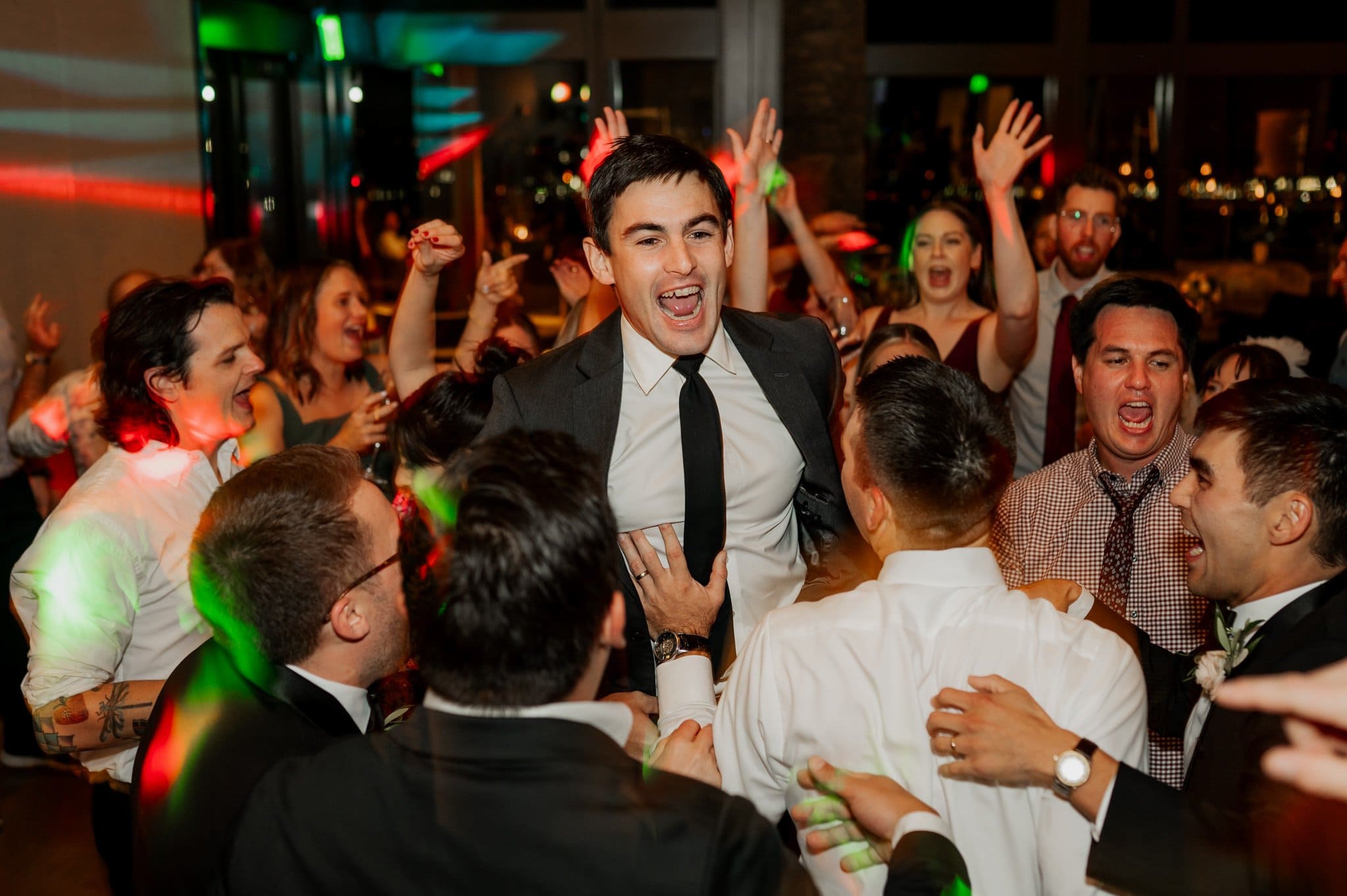 A joyous crowd lifts a man in a suit during a lively celebration at the Skyline Ballroom, part of the renowned Amaterra Winery—a popular Portland wedding venue.