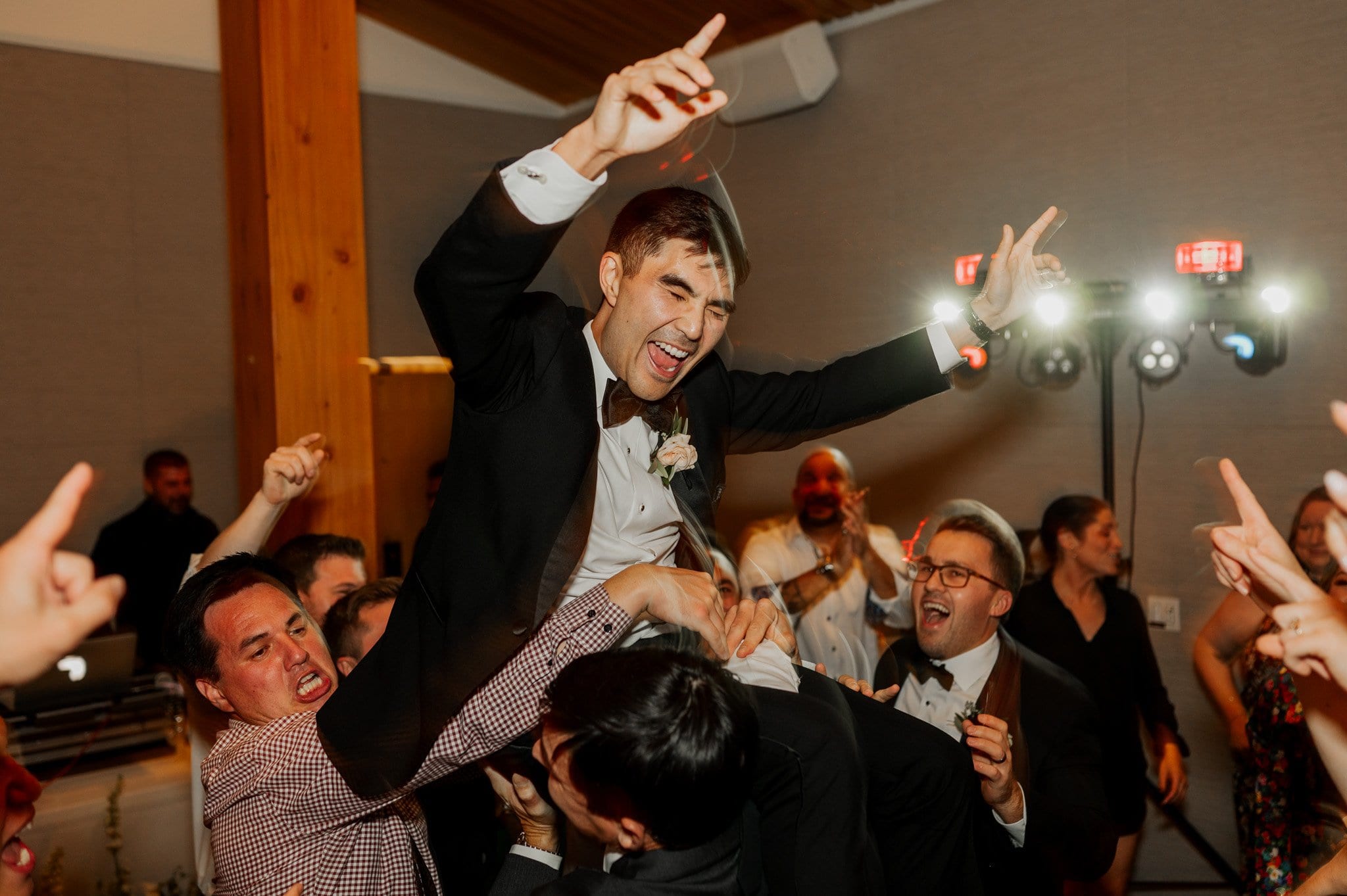 A joyful groom in formal wear is lifted by cheering people at a lively indoor celebration in the Skyline Ballroom of Amaterra Winery, a renowned Portland wedding venue.