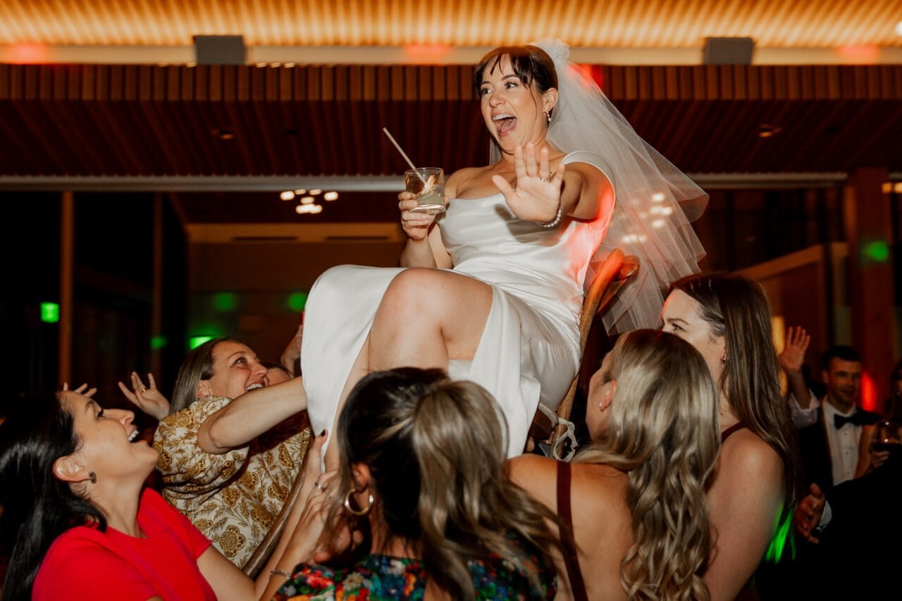 At an Amaterra Winery celebration, the bride in a white dress and veil is joyfully lifted on a chair by guests, smiling as she raises her drink.