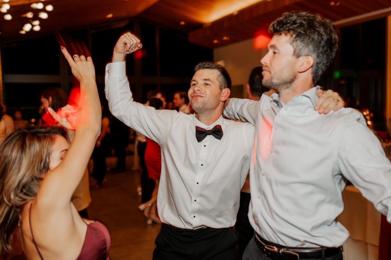 Three people dancing; one flexes muscles. Captured in Amaterra Winery wedding photos, theyre dressed in formal attire amid a festive atmosphere with soft lighting.